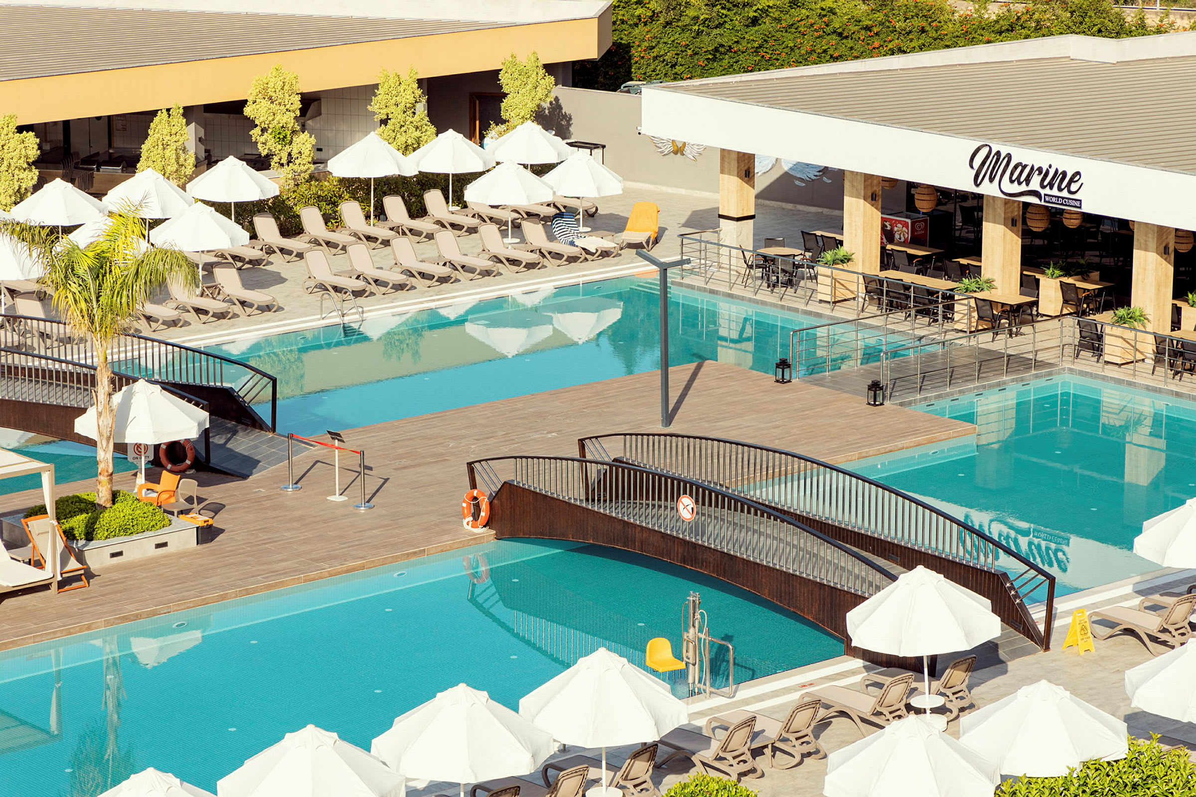Poolområde med liggestole, parasoller, en gangbro over poolen og en restaurant med siddepladser. (AI-genereret tekst)