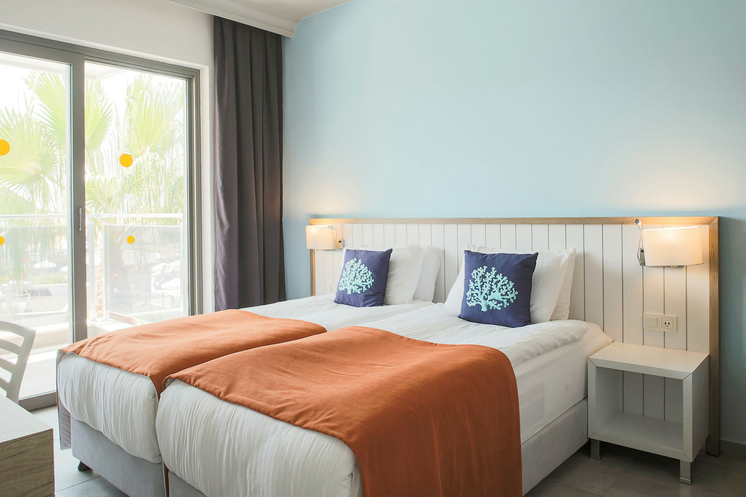 Hotelværelse med to enkeltsenge, orange tæpper og dekorative puder. (AI-genereret tekst)