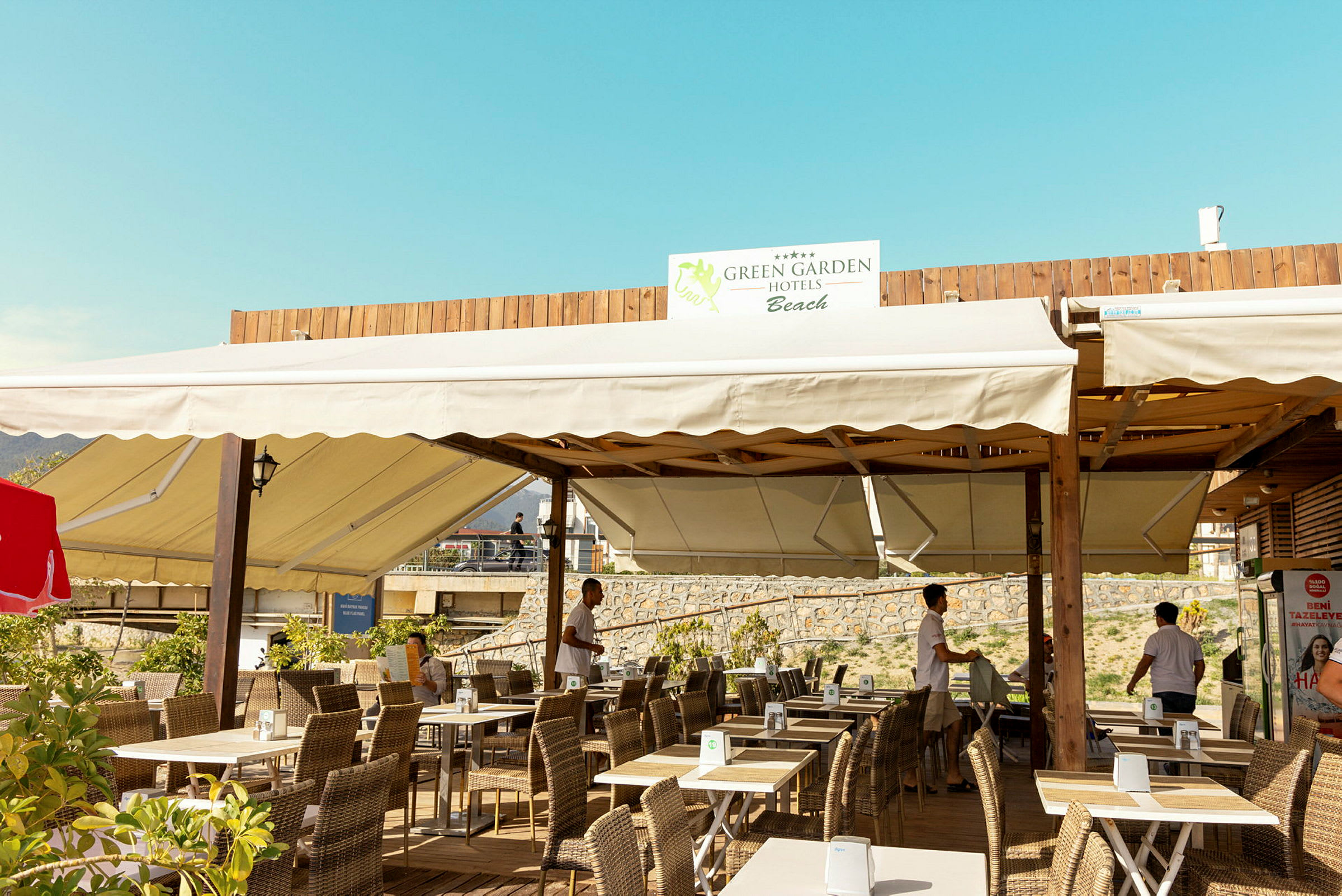 En udendørs servering med borde og stole, med ovenliggende sejltag og et skilt med teksten 'Green Garden Hotels Beach'. (AI-genereret tekst)