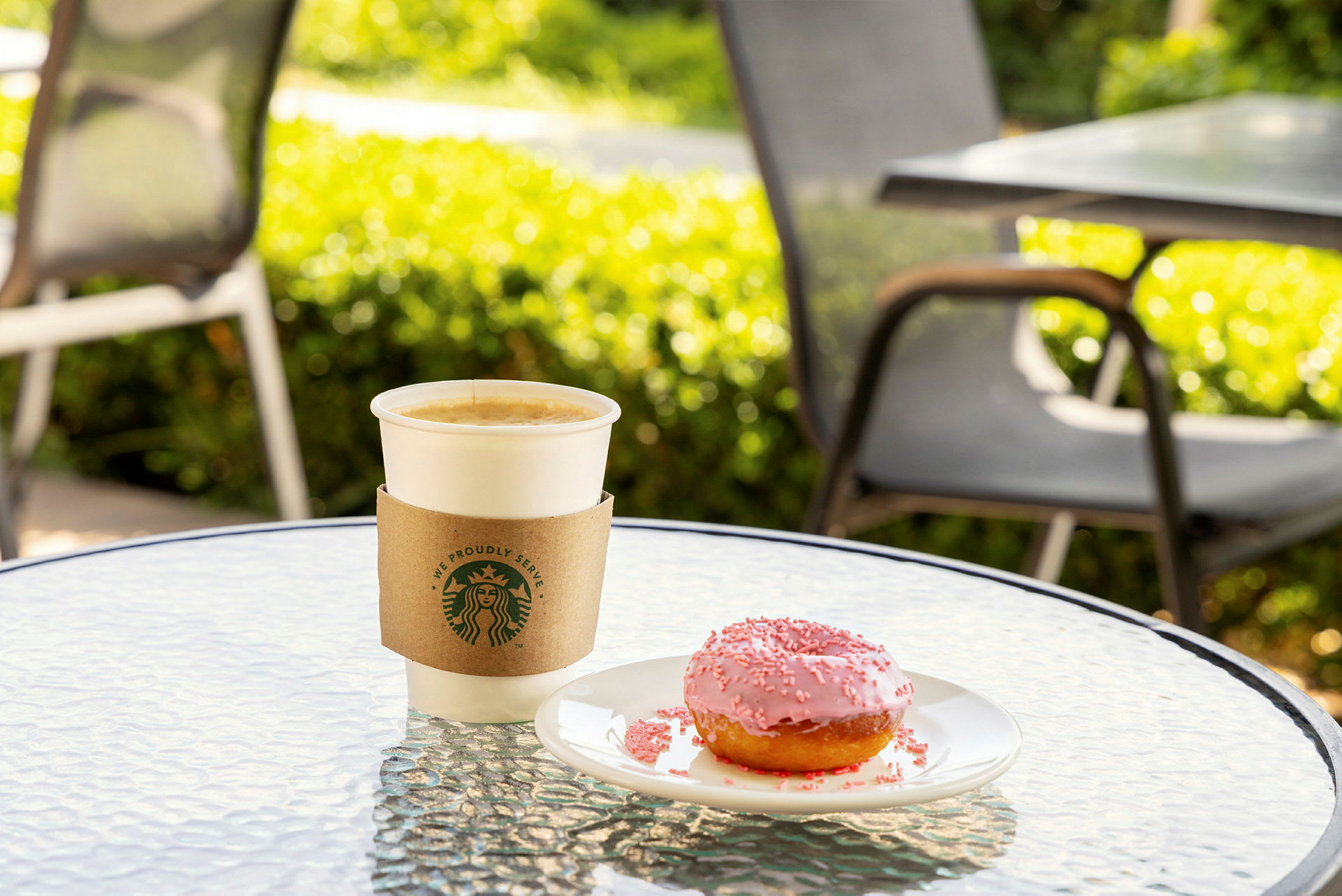En kop kaffe og en lyserød donut på et bord udenfor. (AI-genereret tekst)