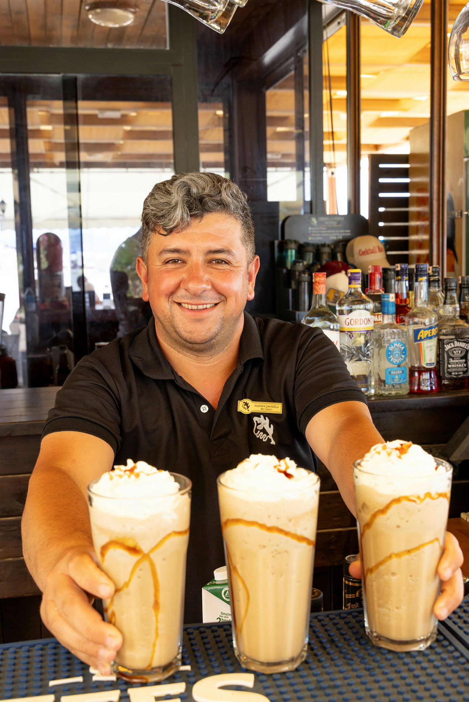 En bartender holder tre glas med milkshakes toppet med flødeskum og karamelsauce. (AI-genereret tekst)