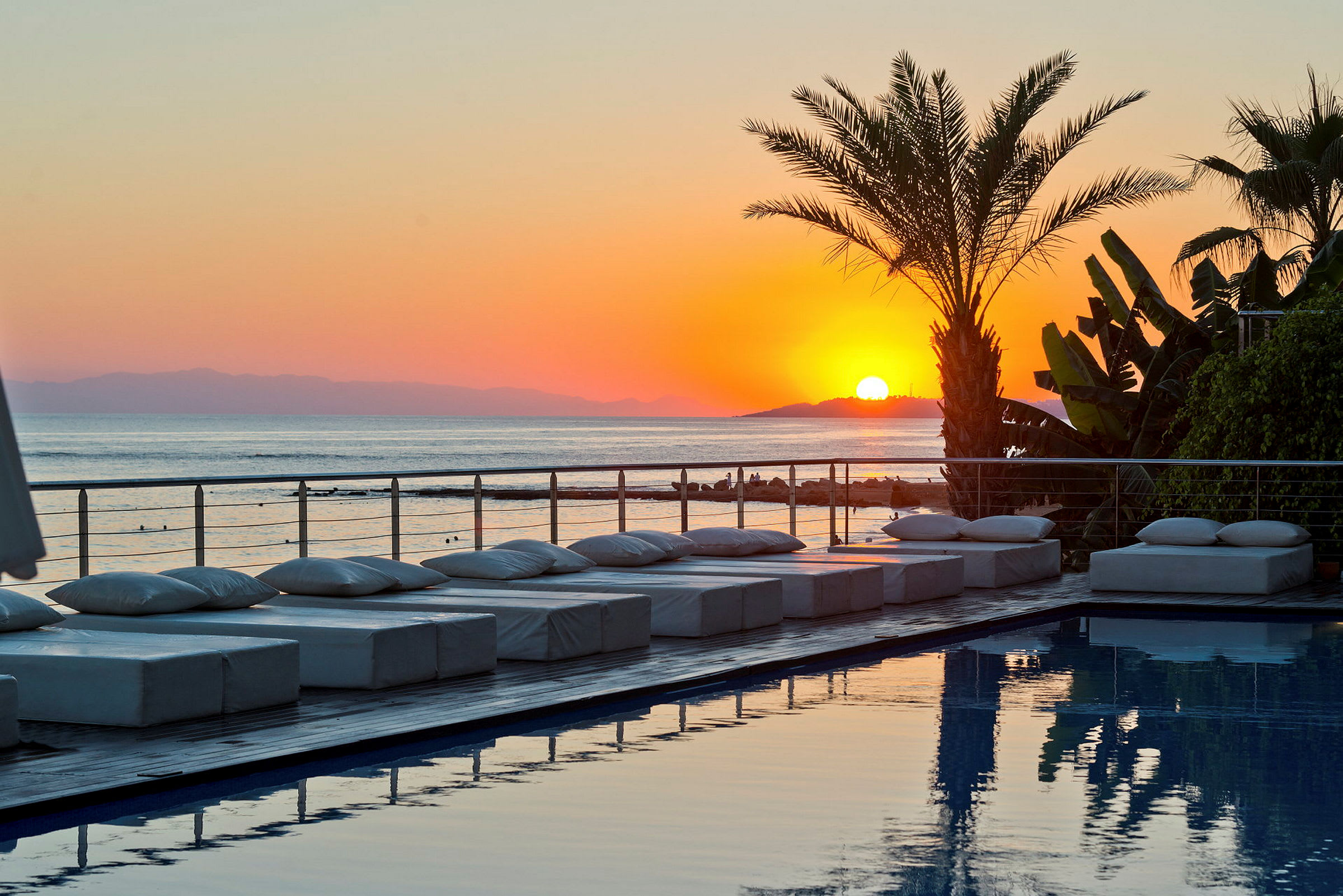 Solnedgang over havet med pool og palmer i forgrunden. (AI-genereret tekst)