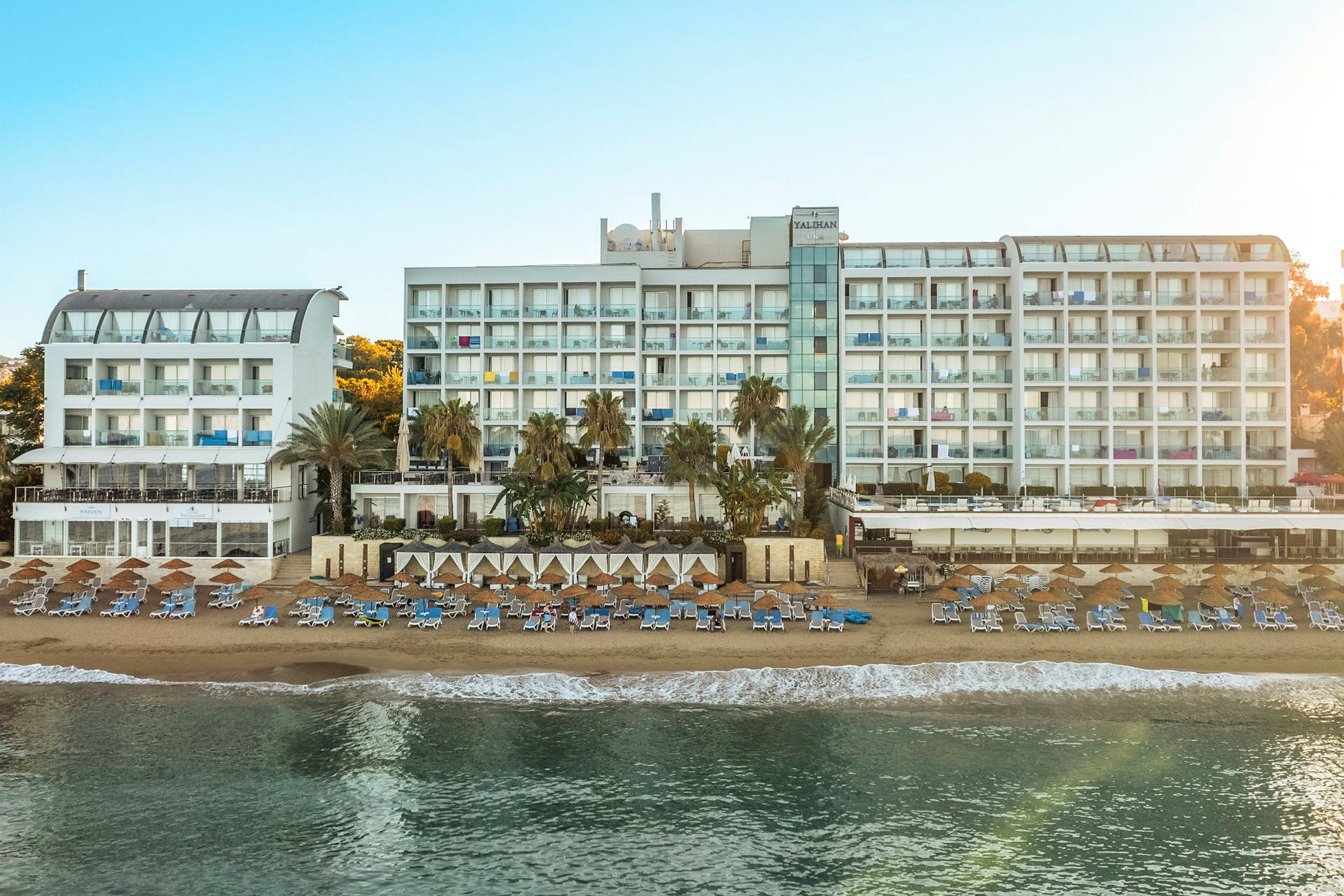 Billede af en stor hvid hotelbygning ved en solrig strand med liggestole og parasoller. (AI-genereret tekst)