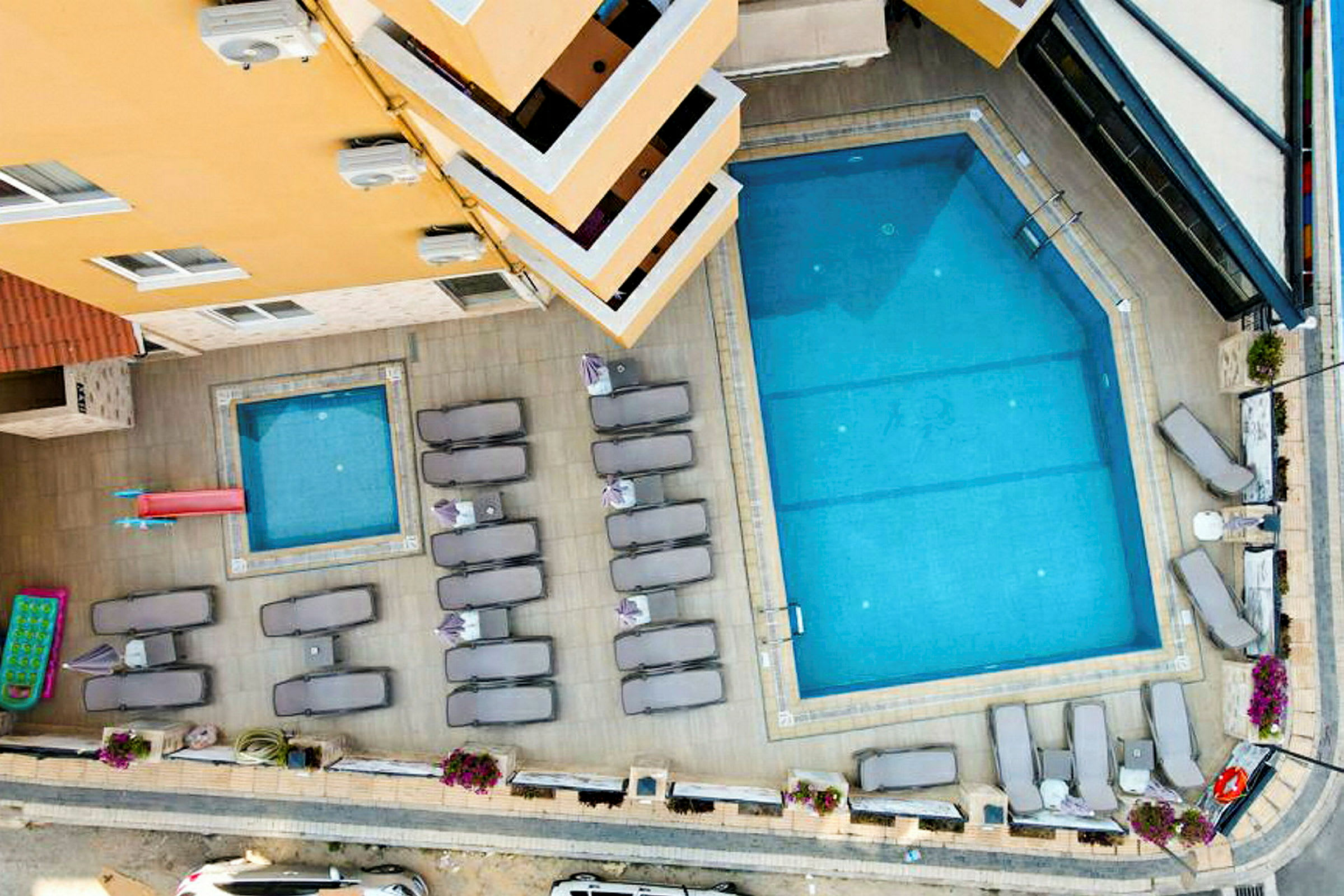 Billede af en udendørs pool og en mindre børnepool, omgivet af liggestole og parasoller ved et hotel. I højre side ses en vej. (AI-genereret tekst)