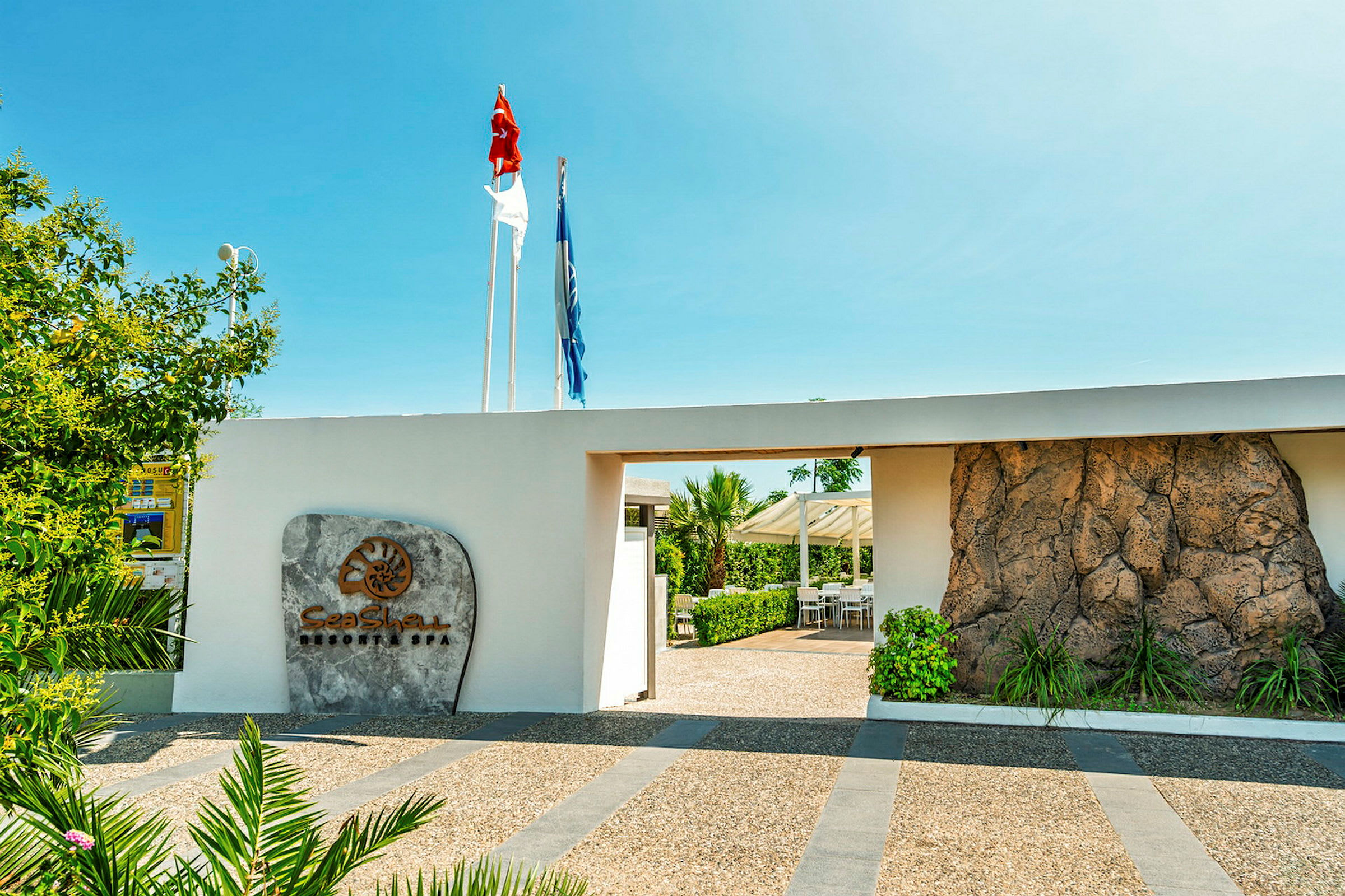 Indgang til en resort med et skilt, der viser navnet 'Sea Shell Resort & Spa', omgivet af grønne omgivelser og tydelige flag. (AI-genereret tekst)