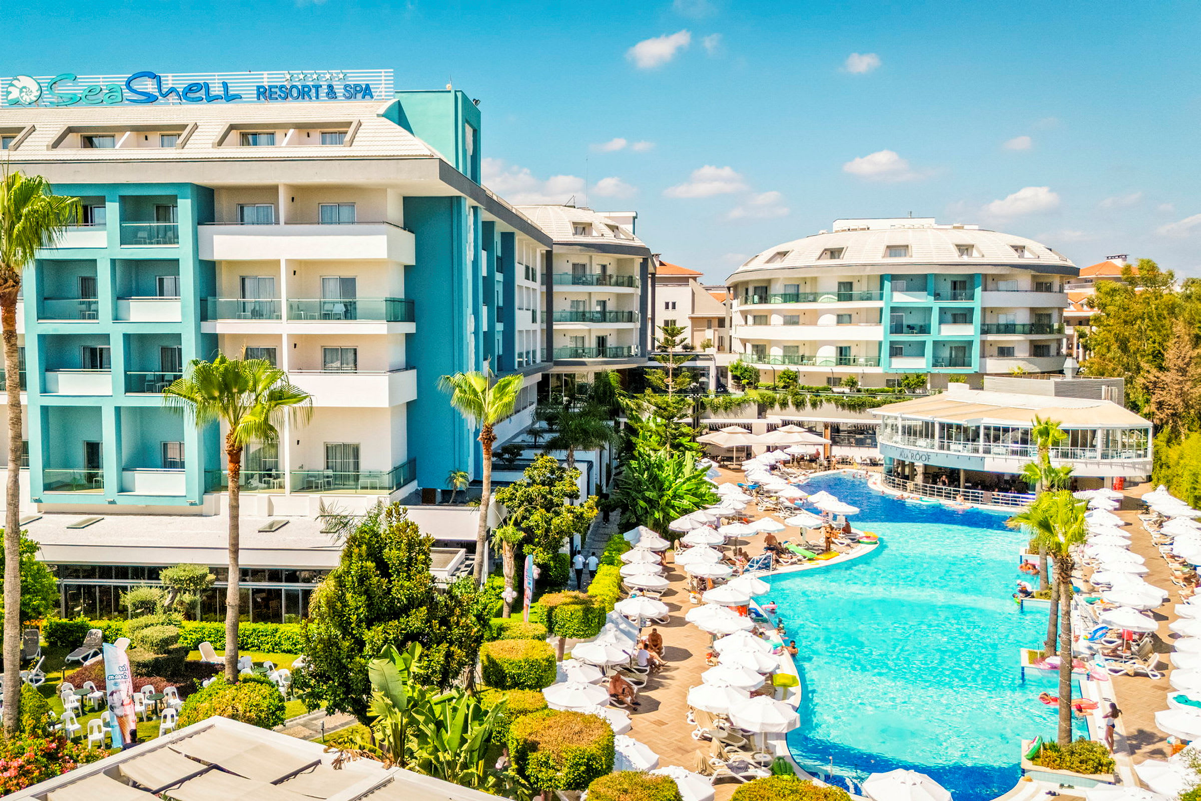 Et billede af et hotel med en stor swimmingpool og flere solsenge. Hotellet har en moderne arkitektur og er omgivet af grønne områder. (AI-genereret tekst)