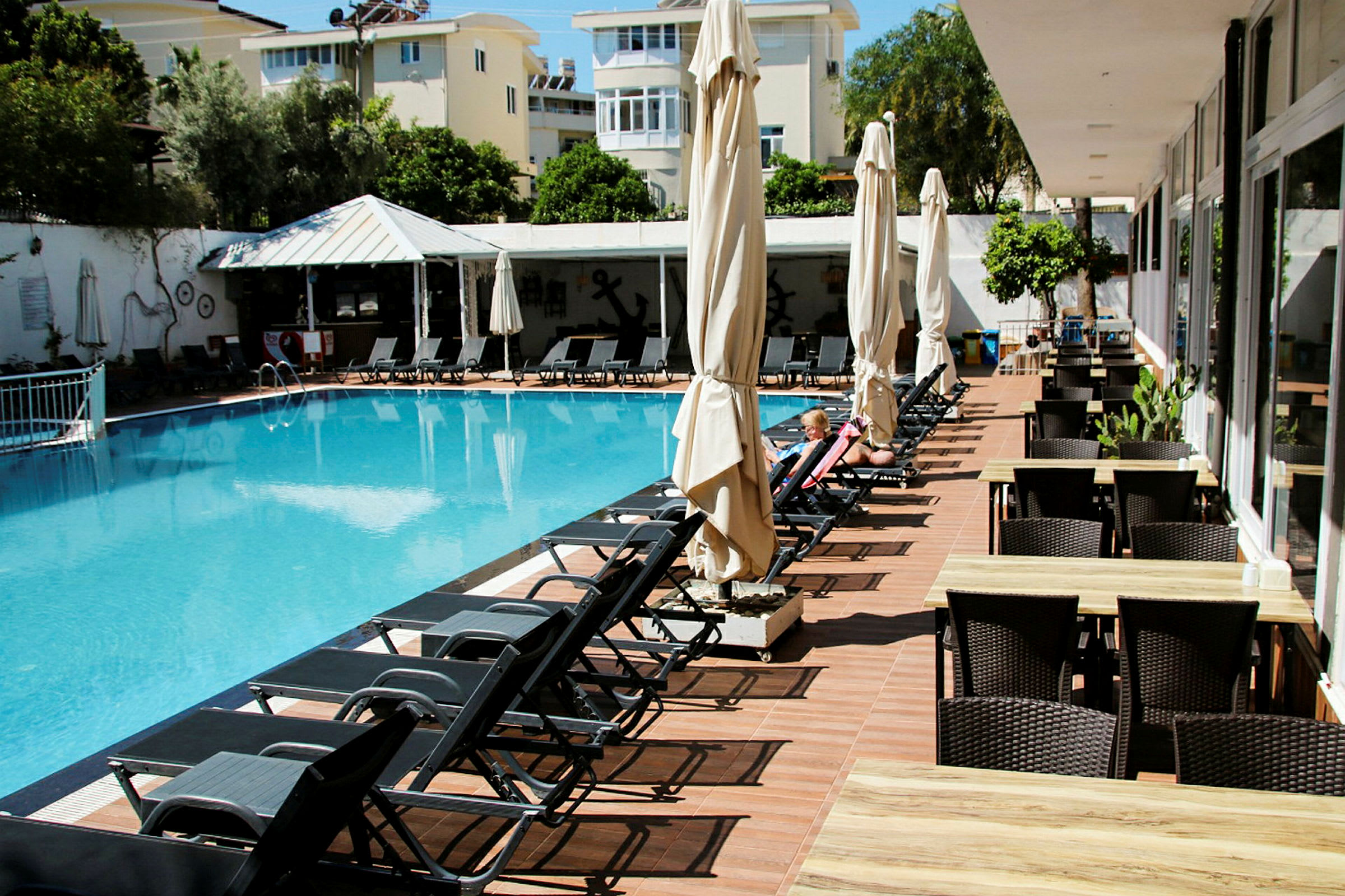 En udendørs swimmingpool med liggestole omkring kanten og borde og stole på en terrasse. (AI-genereret tekst)