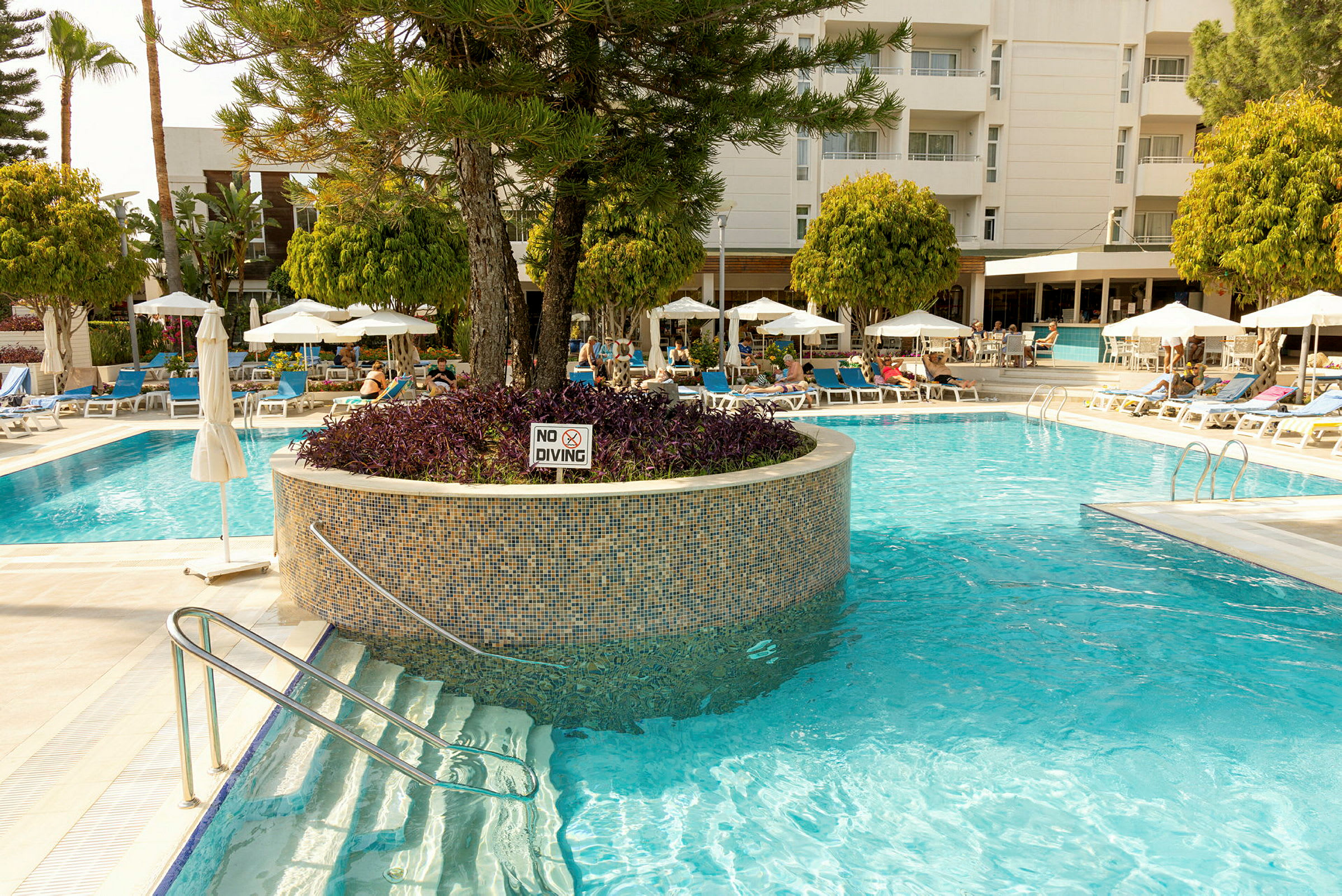 En hotelpool omgivet af solsenge og parasoller. (AI-genereret tekst)