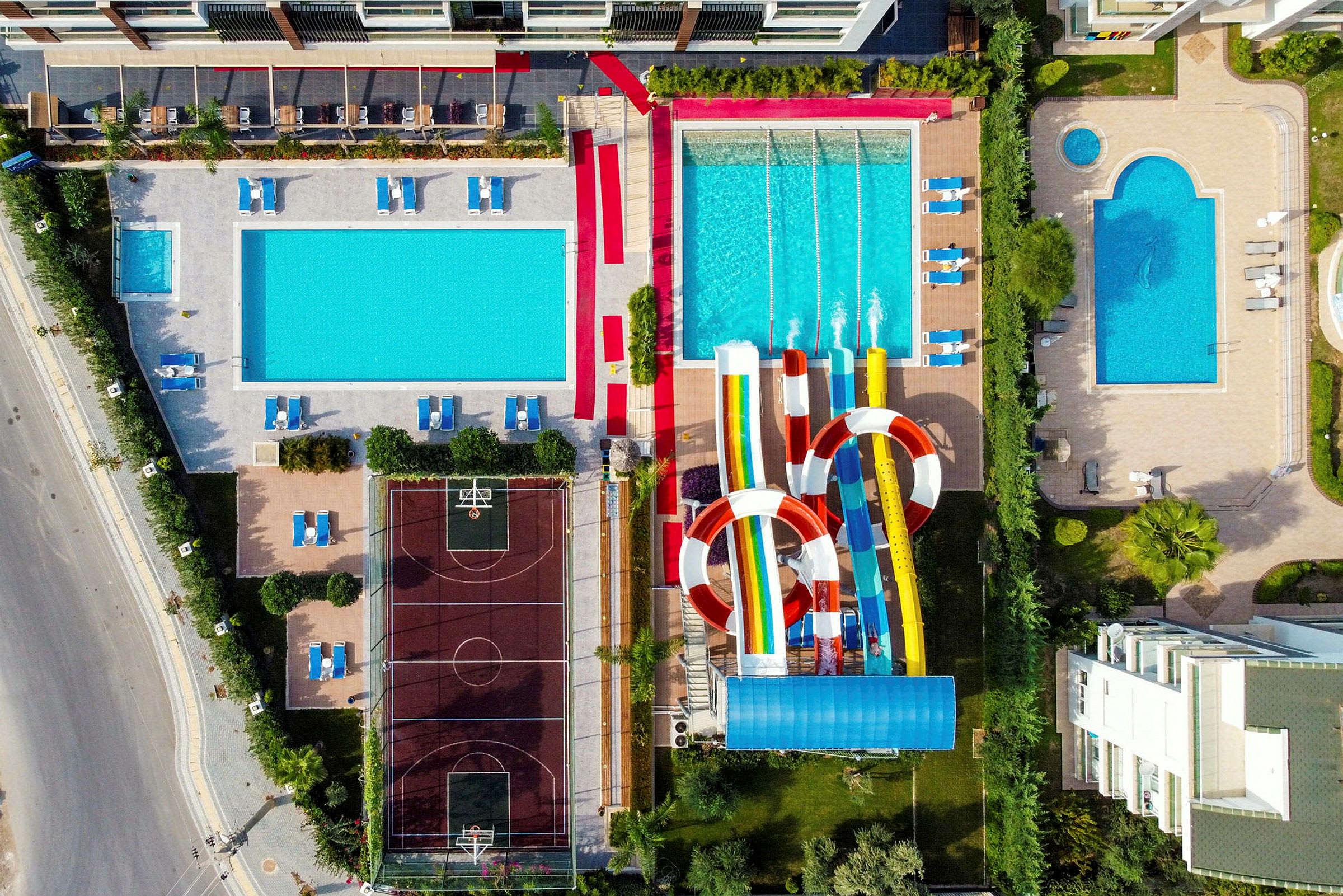 Et fugleperspektivsbillede af et hotelområde med flere pooler, liggestole, vandrutsjebaner og en basketballbane. (AI-genereret tekst)
