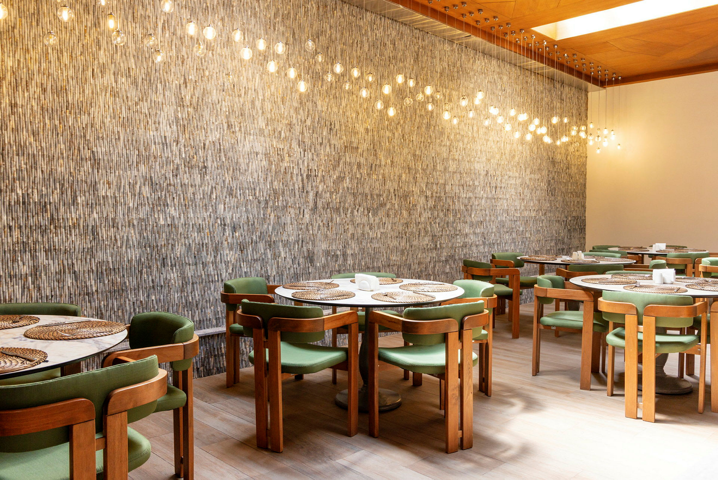 En indendørs restaurant med træstole og grønne sæder, runde borde med sivmåtter og hængende lamper med glødepærer over. (AI-genereret tekst)