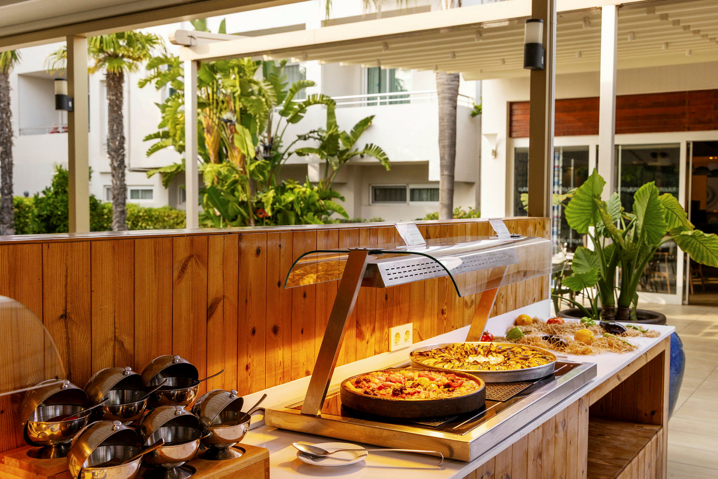 En buffet med mad, inklusive to pizzaer, omgivet af grønne planter og et udemiljø. (AI-genereret tekst)