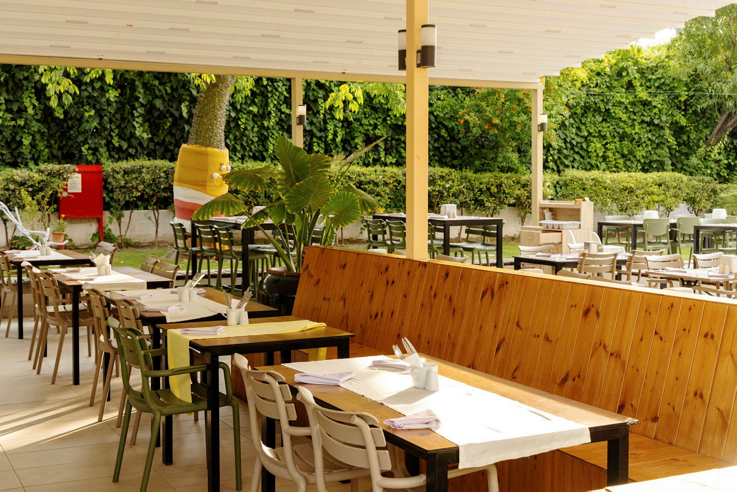 En udendørsrestaurant med træborde, stole og en stor grøn plante i et havemiljø. (AI-genereret tekst)