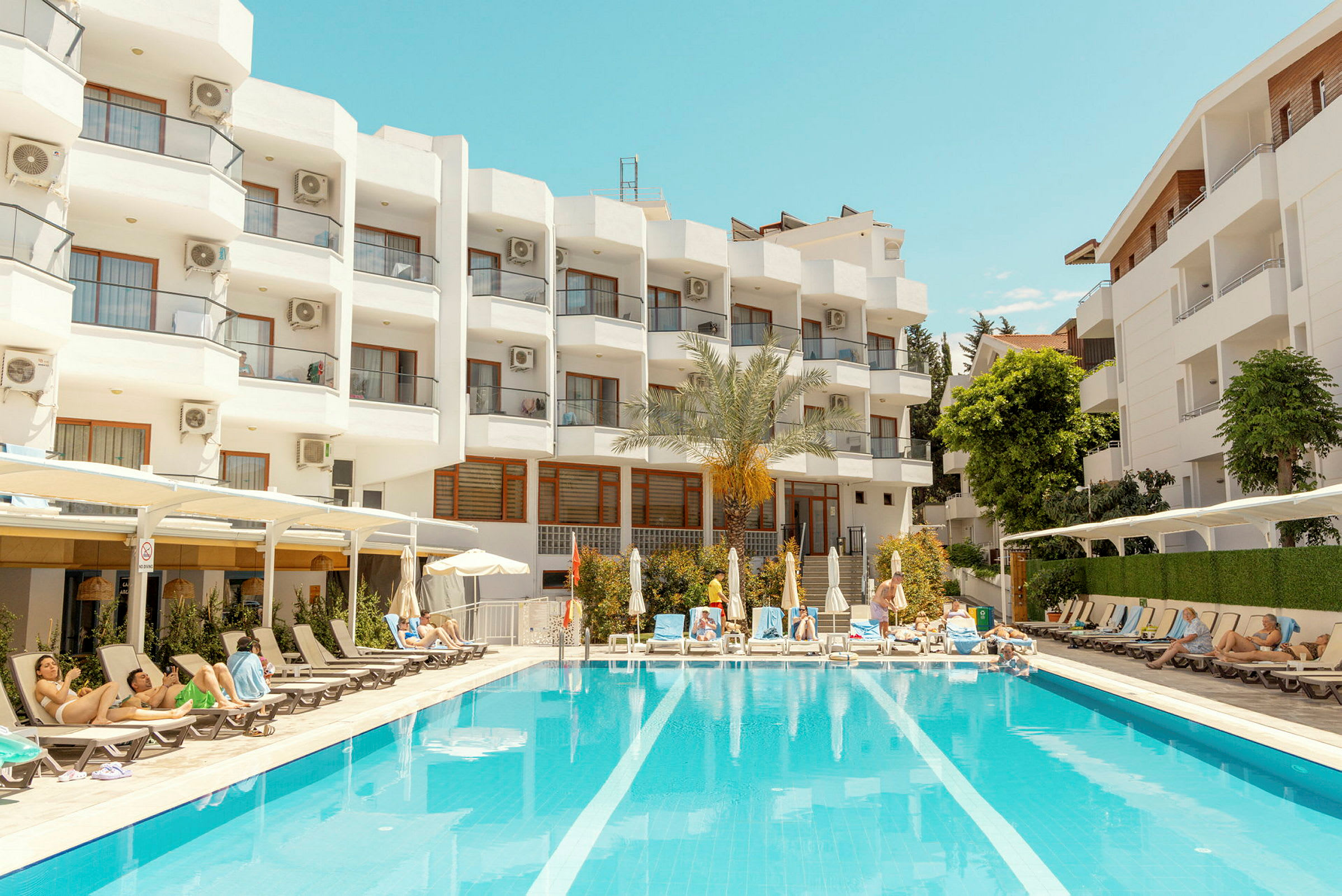 Et hotel med hvid facade og altaner ses i baggrunden. Foran bygningen ligger en udendørspool omgivet af solsenge og parasoller, hvor folk slapper af. (AI-genereret tekst)