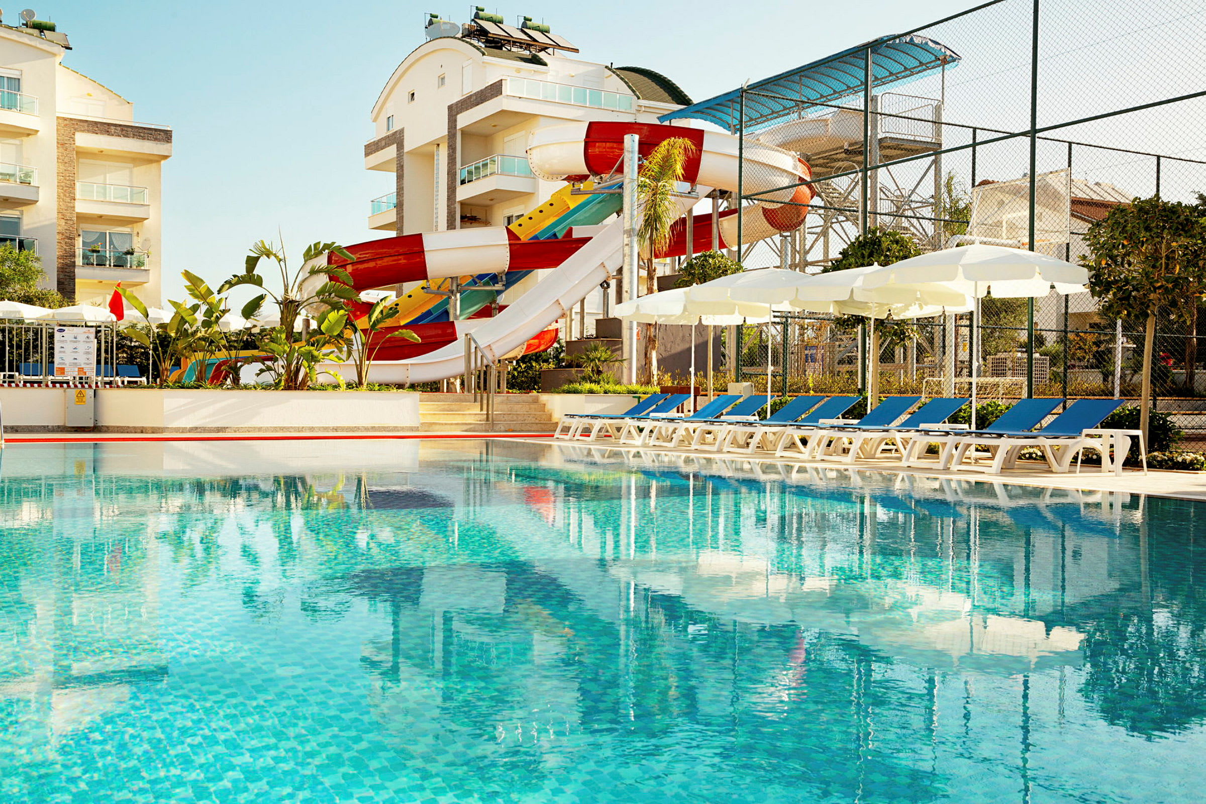 Udendørs swimmingpool med solsenge, parasoller og en farverig vandrutsjebane ved siden af moderne bygninger. (AI-genereret tekst)