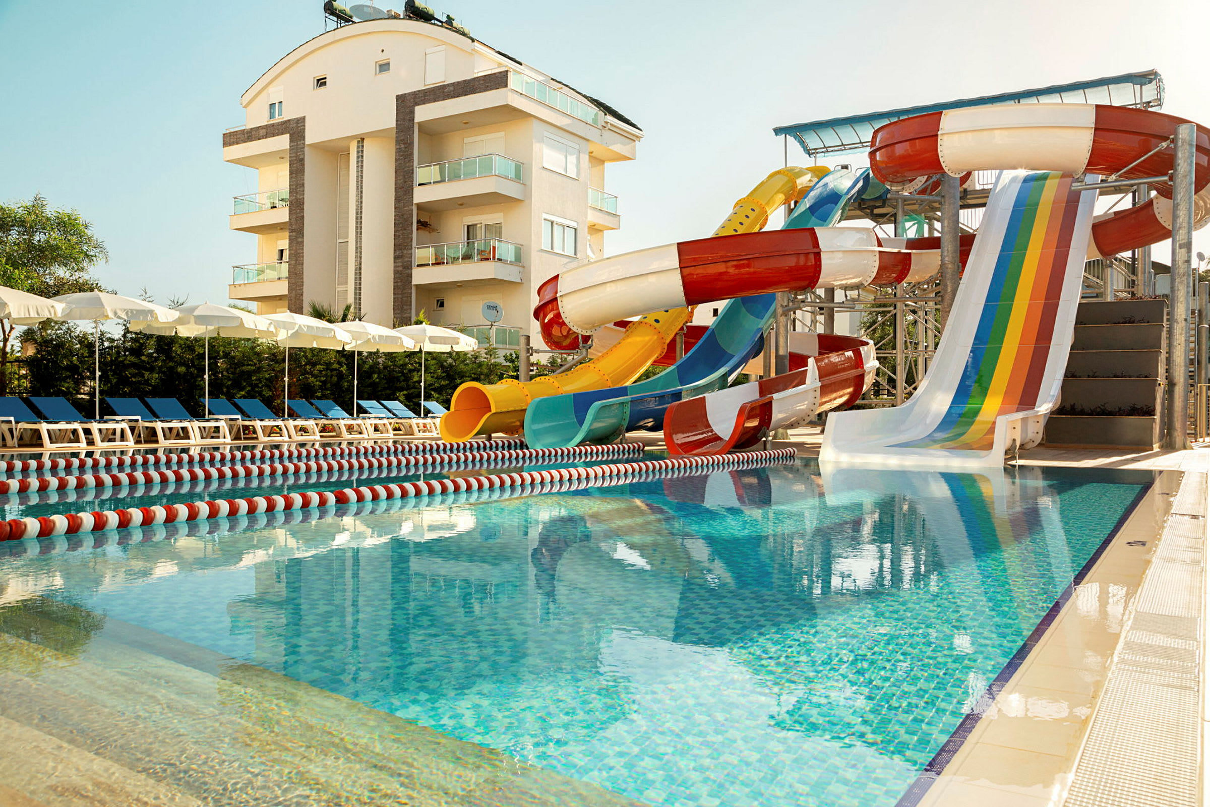 Udendørs swimmingpool med vandrutsjebaner og liggestole nær en lejlighedsbygning. (AI-genereret tekst)