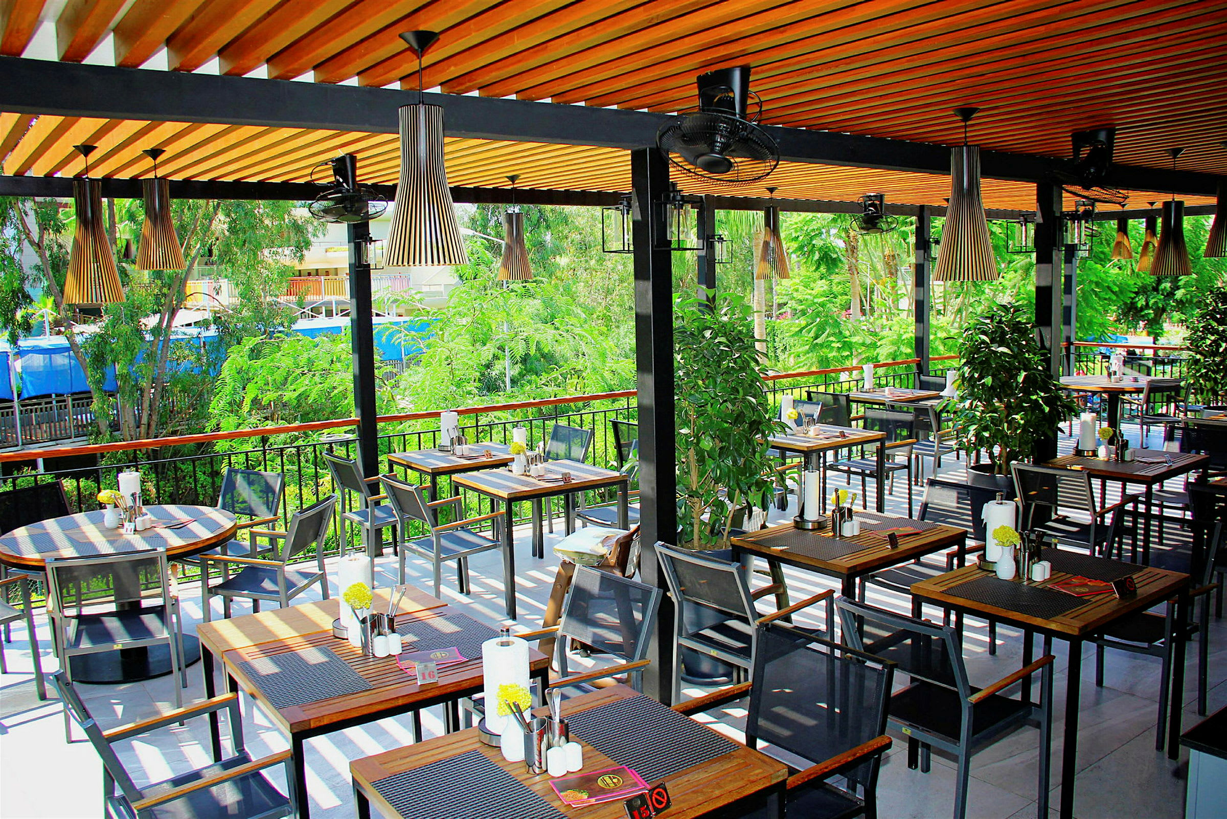 Udendørs restaurant med borde og stole, omgivet af grønne områder. (AI-genereret tekst)