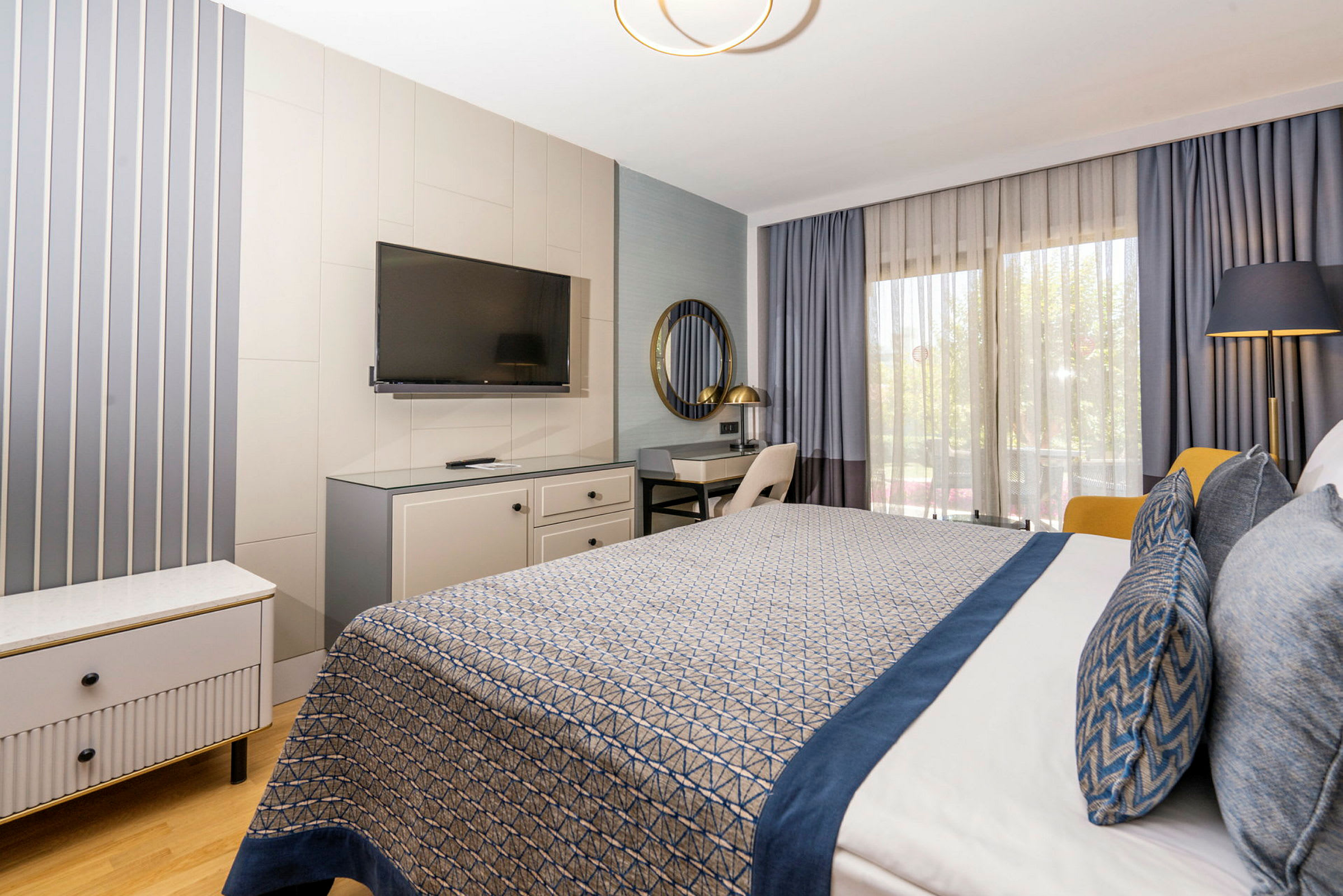 Hotelværelse med moderne indretning, inklusive seng med blå detaljer, skrivebord, TV og store vinduer med gardiner. (AI-genereret tekst)