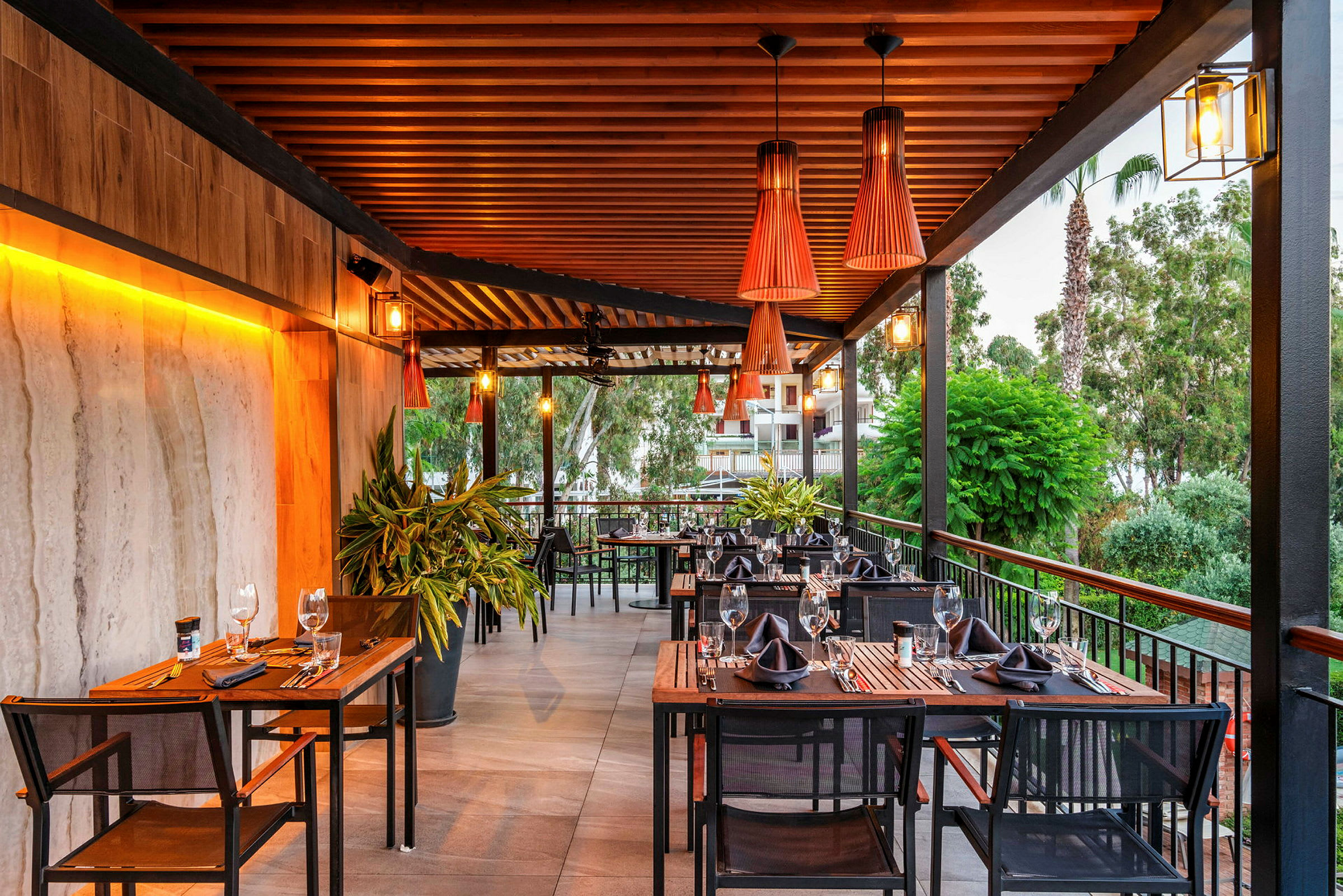 En terrasse med borde og stole arrangeret til middag, med grønt og dekorative lamper. (AI-genereret tekst)
