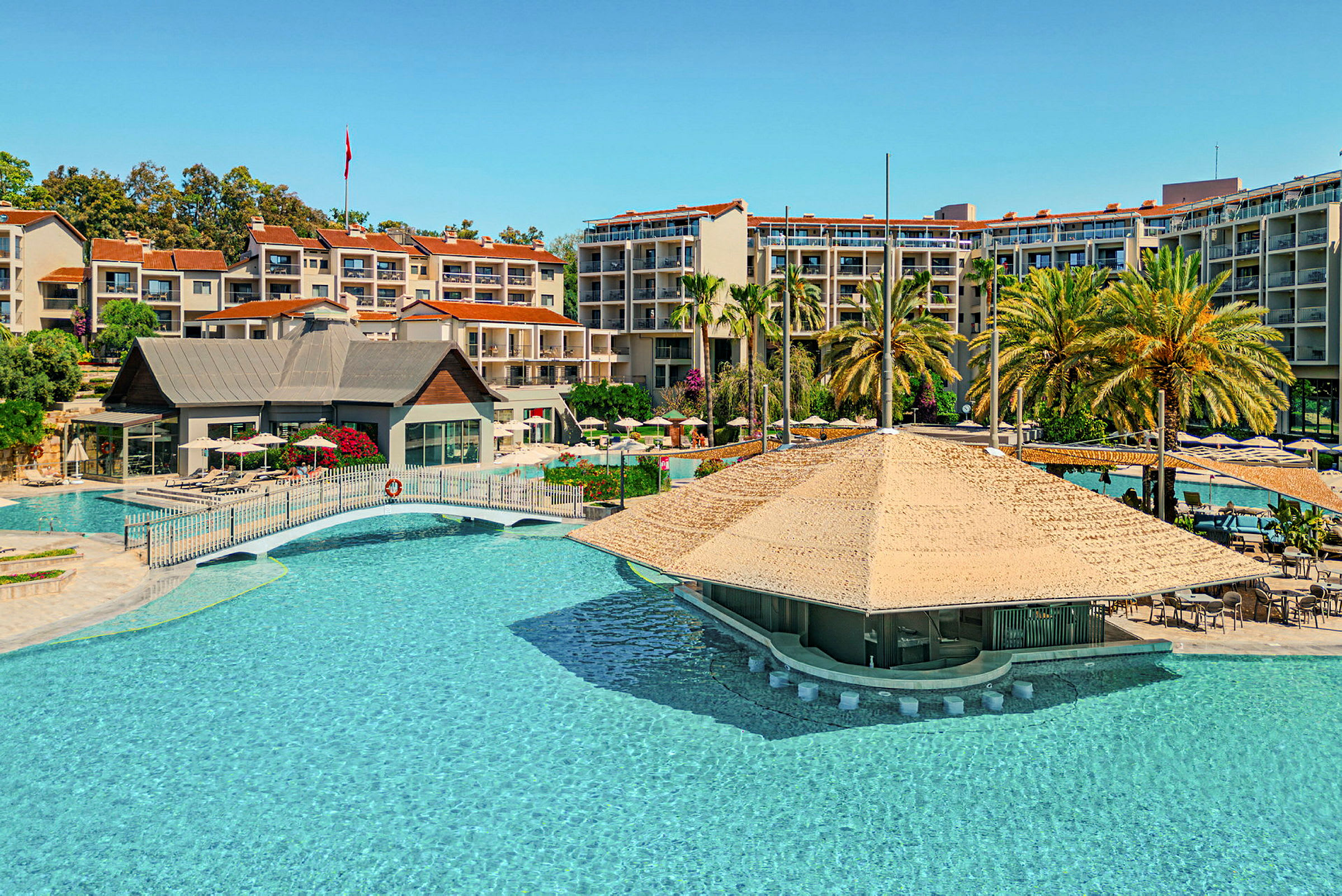 Billede af et hotelområde med en stor swimmingpool, overdækket poolbar og omkringliggende bygninger samt palmer. (AI-genereret tekst)