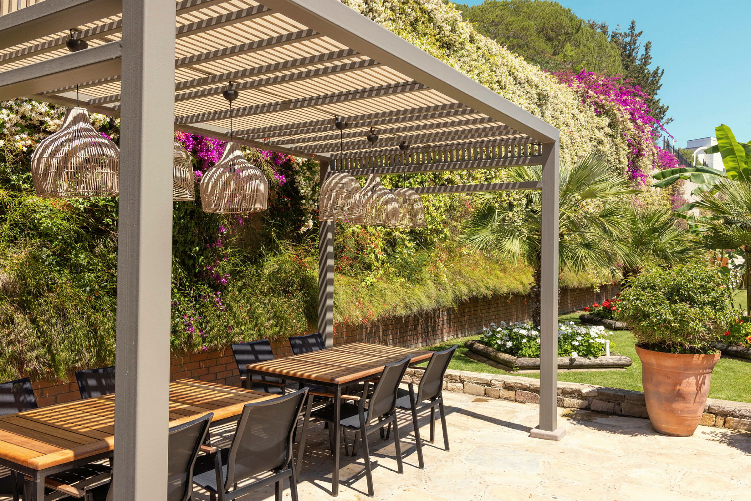 En udendørs siddeområde med borde og stole under en pergola, omgivet af grønt og blomster. (AI-genereret tekst)