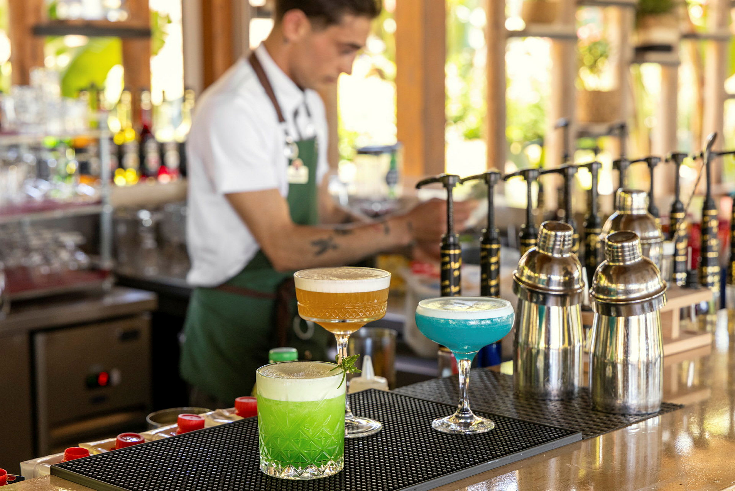 En bar med tre farverige cocktails, shakere og en bartender i baggrunden. (AI-genereret tekst)