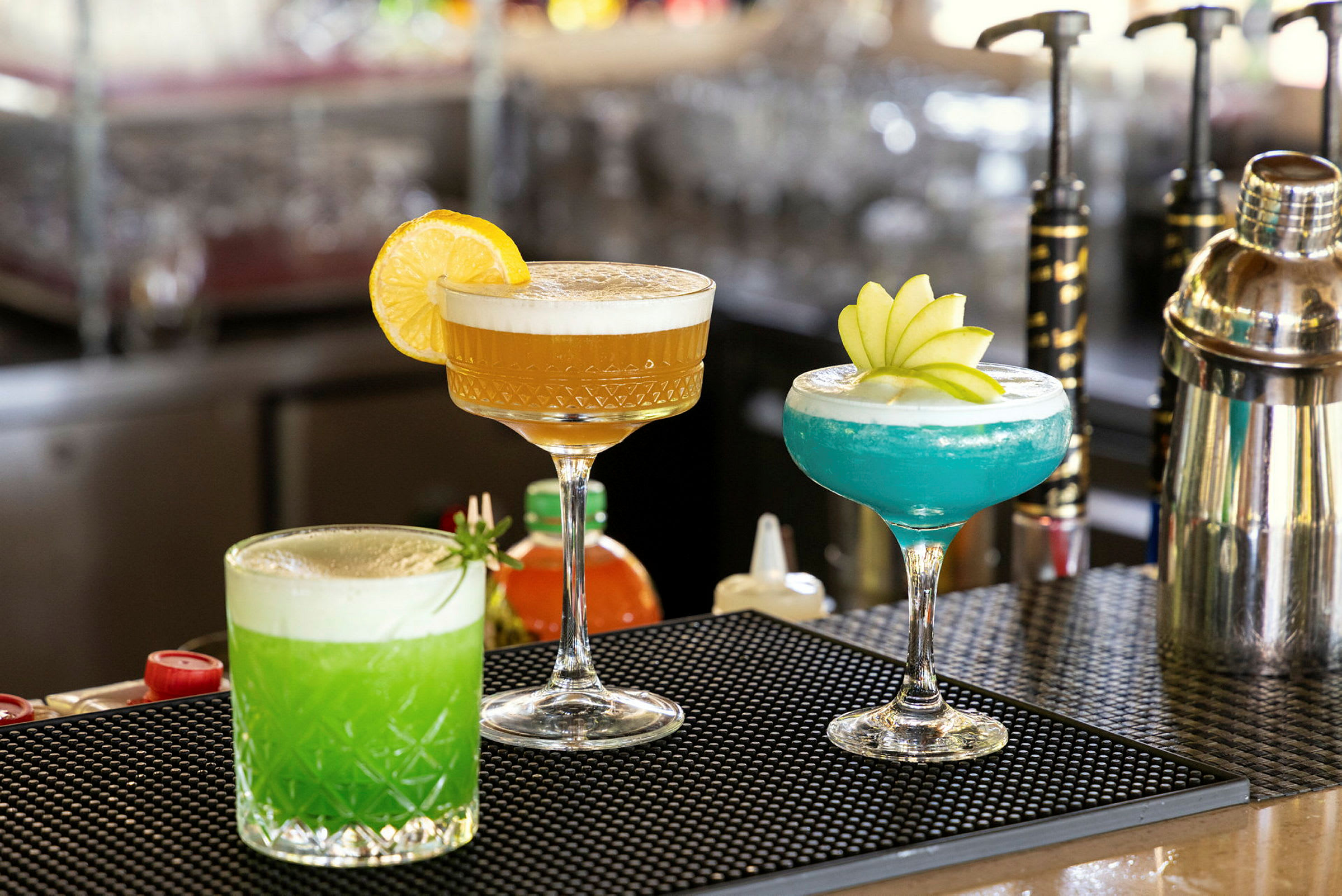 Tre farverige drinks på en bardisk, pyntet med frugt og krydderurter. (AI-genereret tekst)