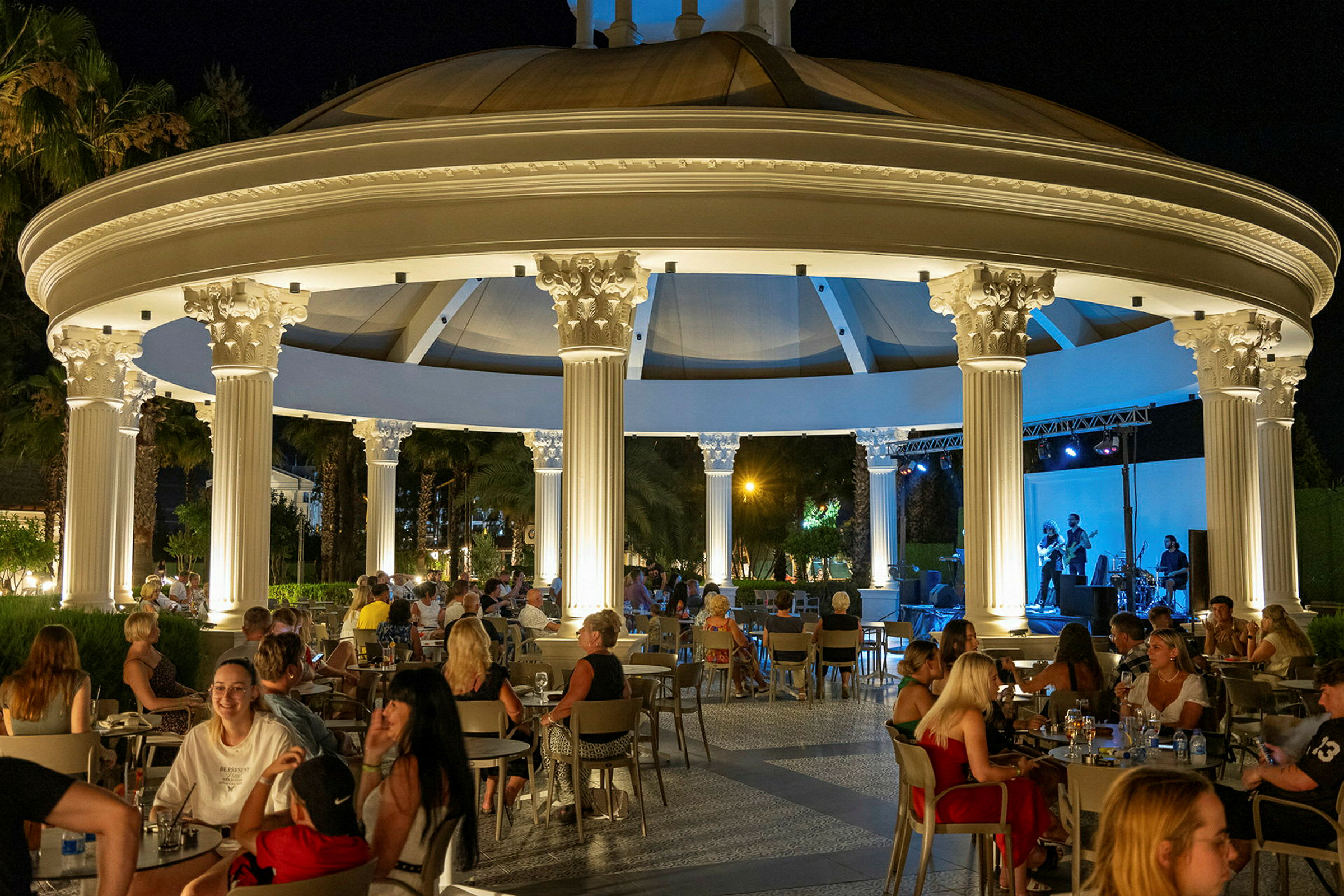 Udendørs restaurant med koncert om aftenen, med belysning og mennesker ved bordene. (AI-genereret tekst)