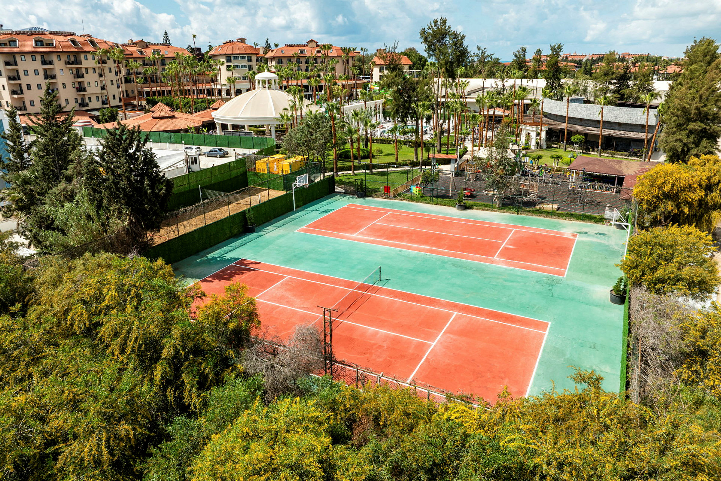 Billede af to tennisbaner omgivet af grønne områder og bygninger i baggrunden. (AI-genereret tekst)