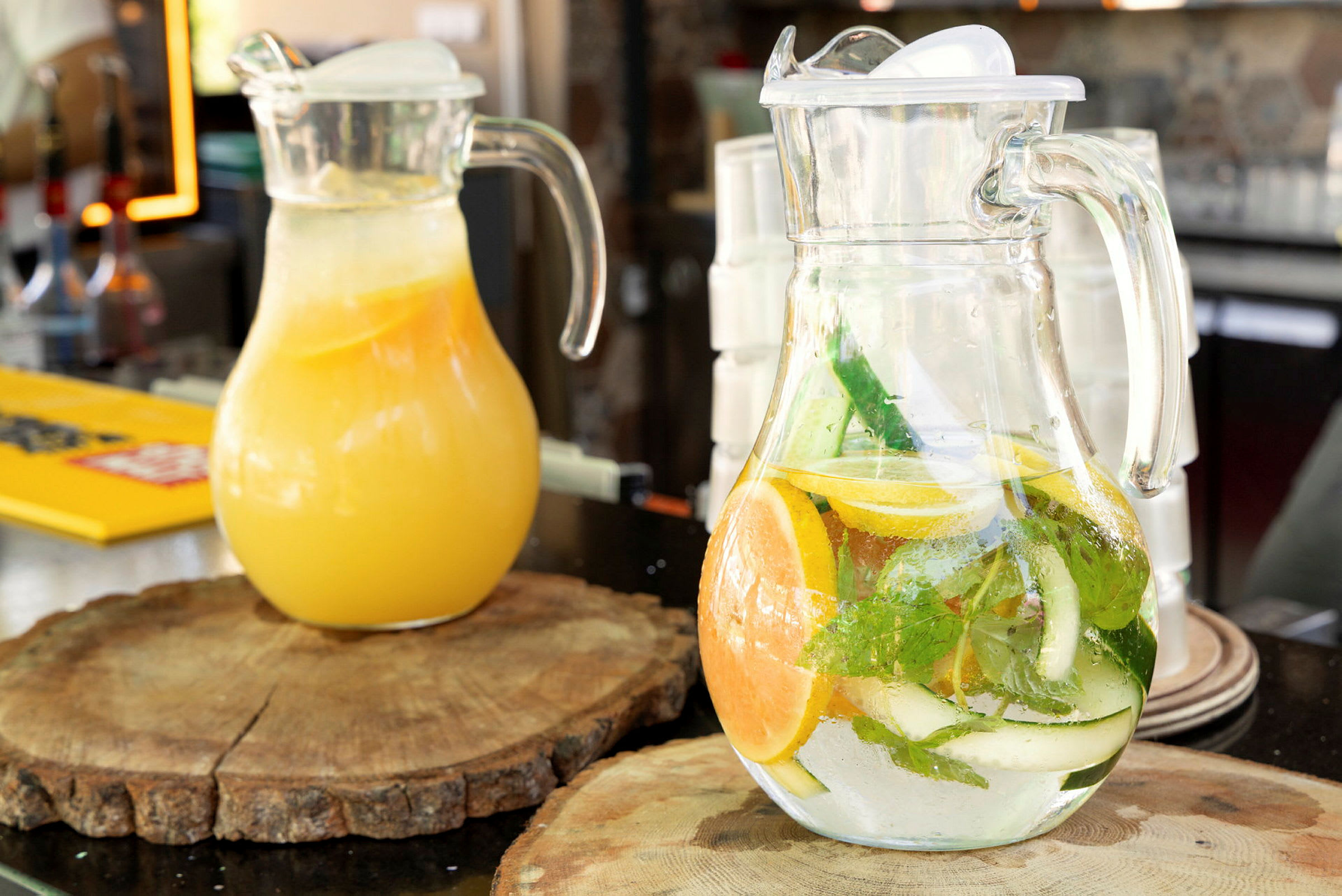 To karafler med drikke, en med appelsinjuice og en med smagstilsat vand med citrus og urter, står på træplader på et bord. (AI-genereret tekst)