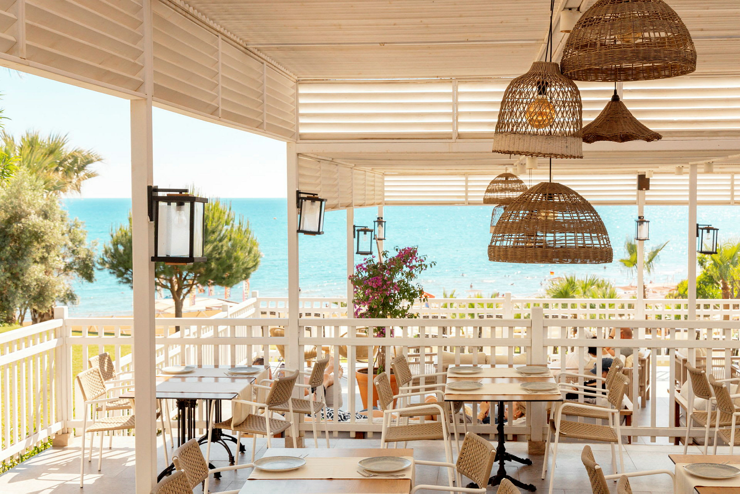 En strandrestaurant med træborde, flettestole og udsigt over havet. (AI-genereret tekst)