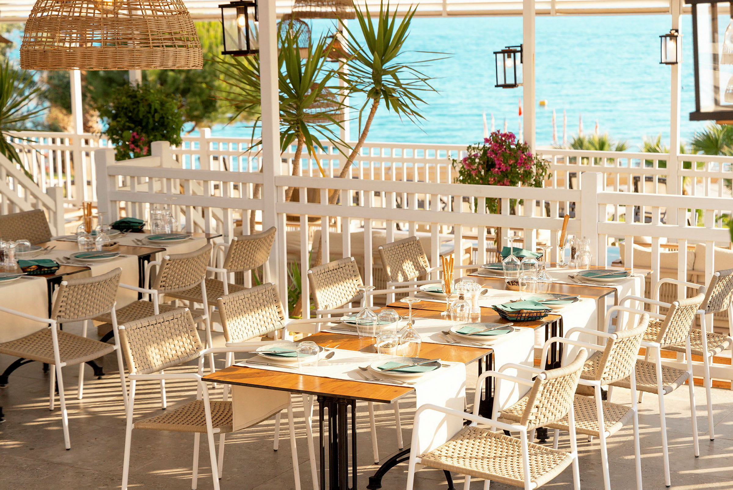 En udendørs restaurant med borde dækket med tallerkener og glas. Udsigt over havet i baggrunden. (AI-genereret tekst)