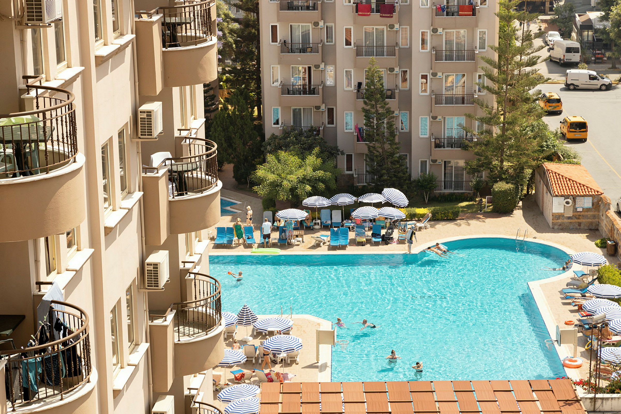 En hotelpool med liggestole og parasoller tæt på bygninger. (AI-genereret tekst)