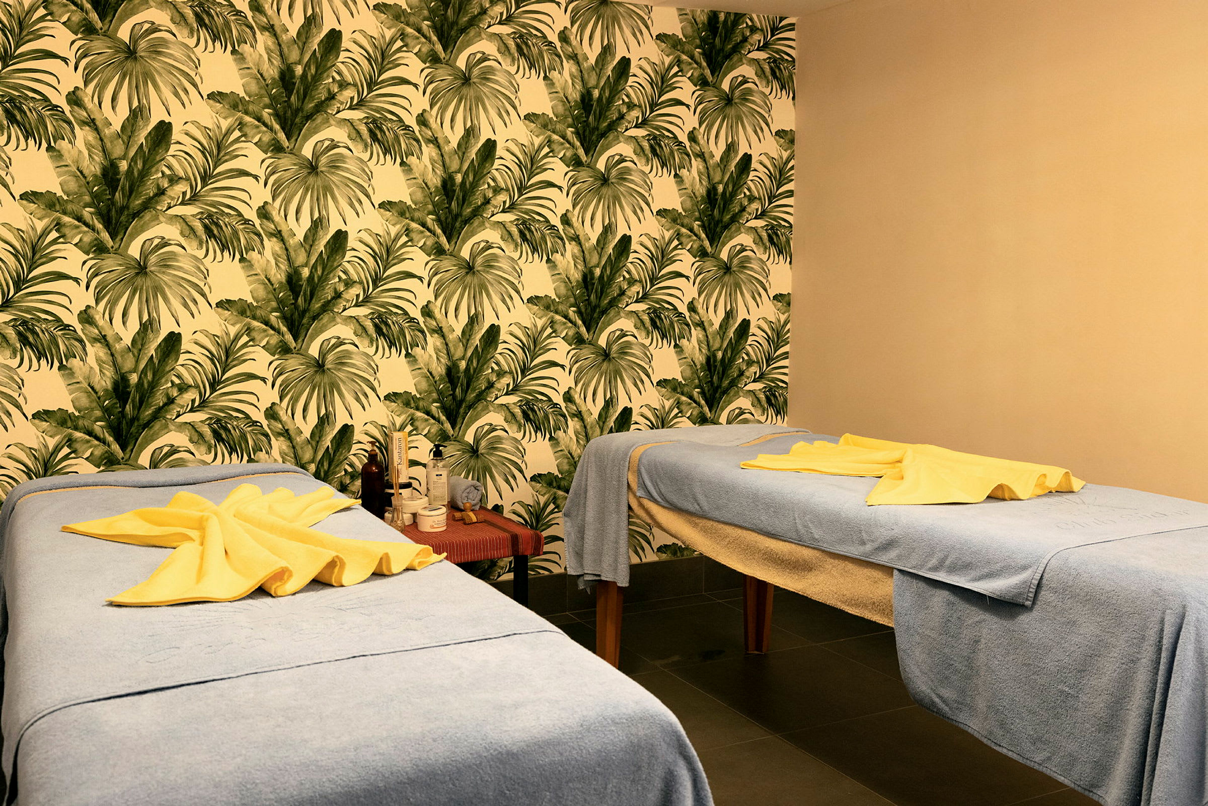 To massageborde med blå håndklæder og gule håndklæder ovenpå i et rum med tropisk bladmotiv på væggen. (AI-genereret tekst)