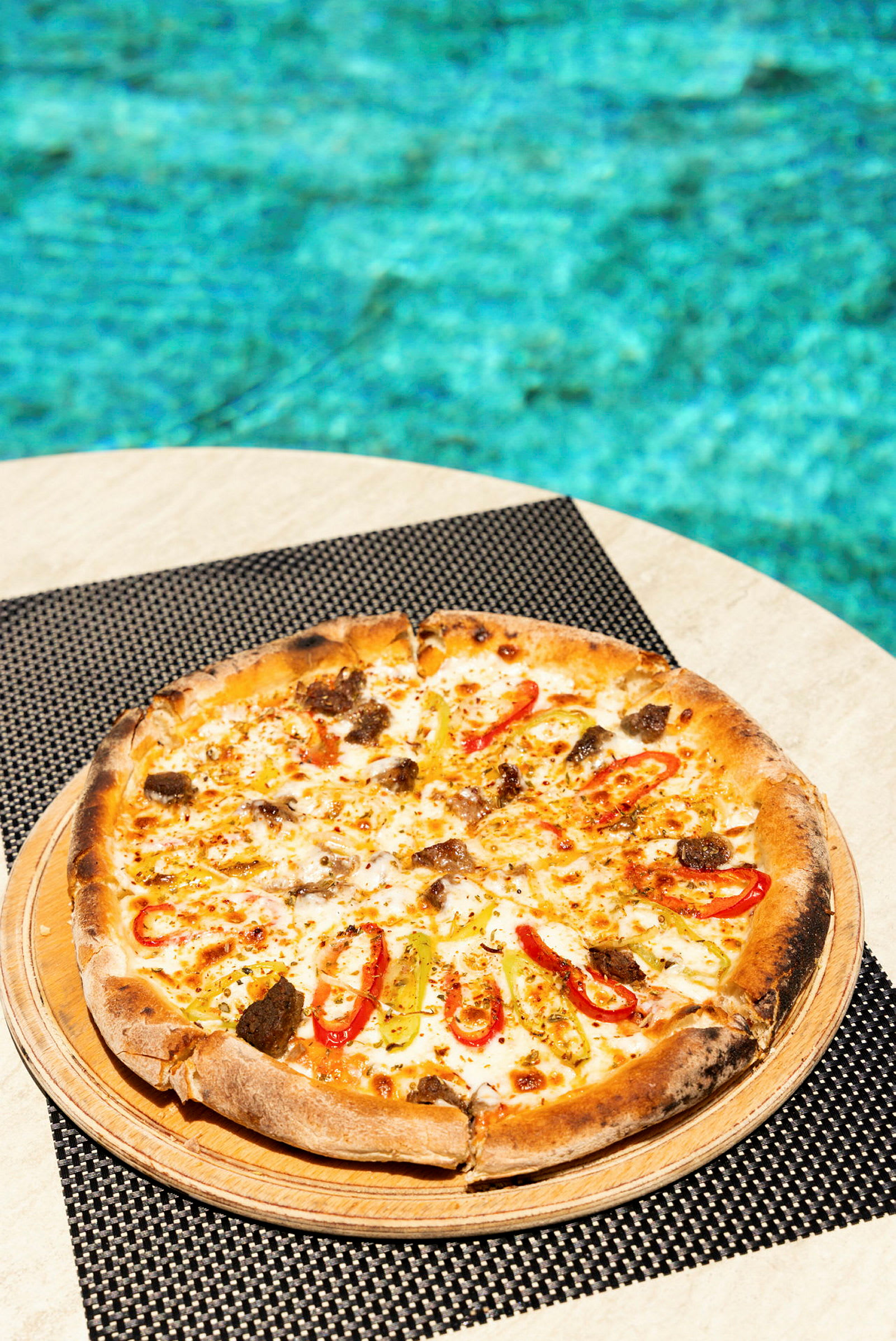 En pizza med fyld som peberfrugtskiver og andet serveres på et bord med en pool i baggrunden. (AI-genereret tekst)