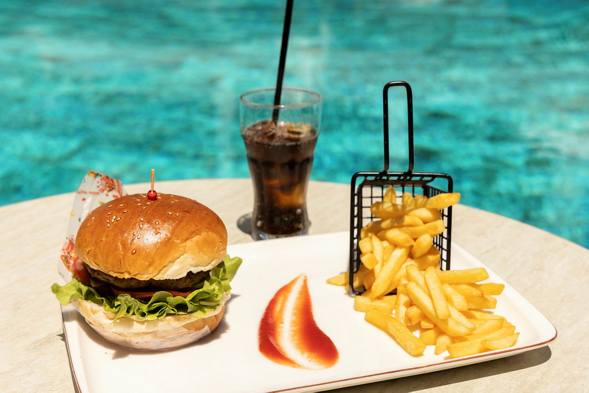 En burger, pommes frites og et glas sodavand på et bord ved en pool. (AI-genereret tekst)