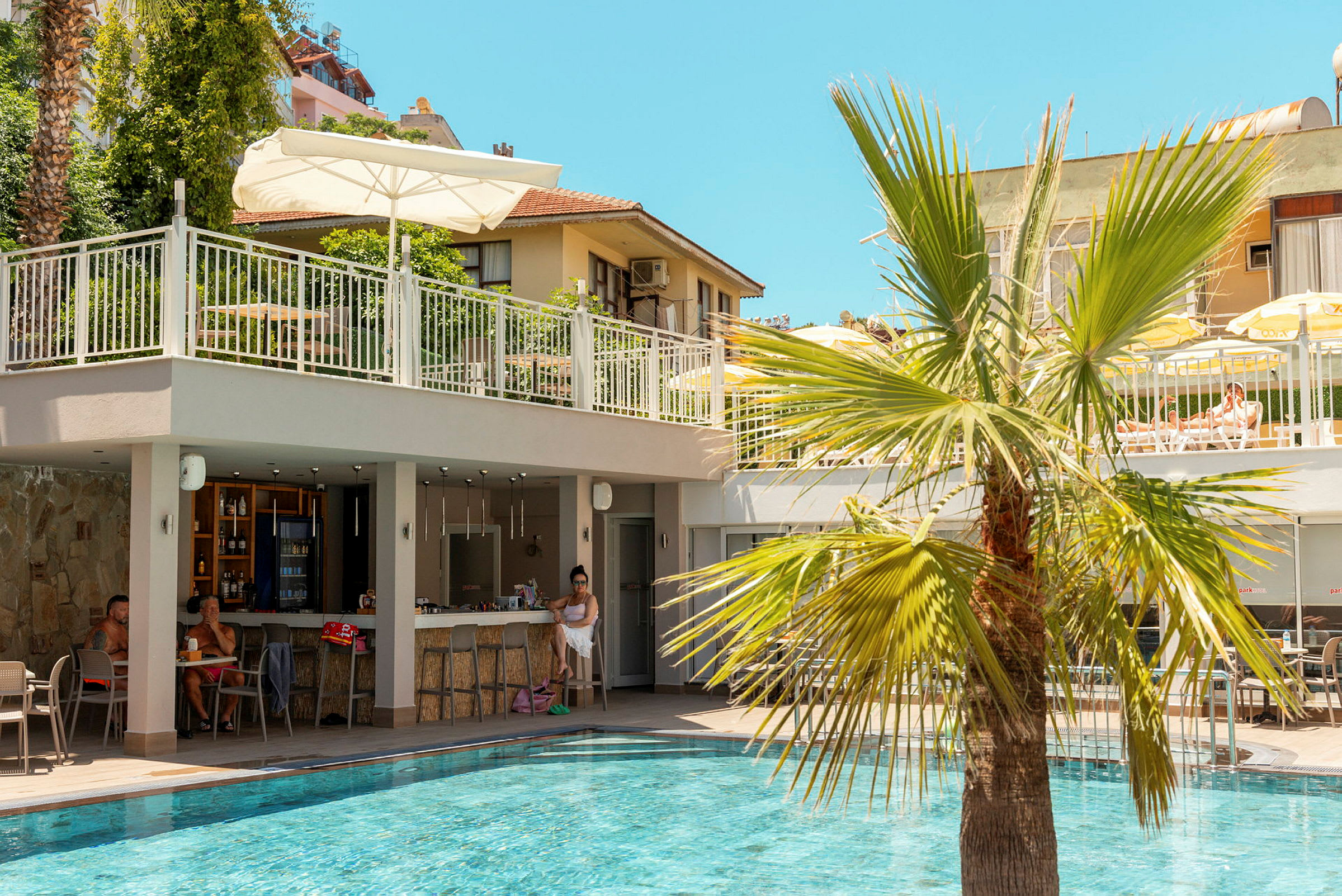 En swimmingpool med en bar ved siden af og en palme i forgrunden. I baggrunden ses en bygning og en terrasse med borde og parasoller. (AI-genereret tekst)