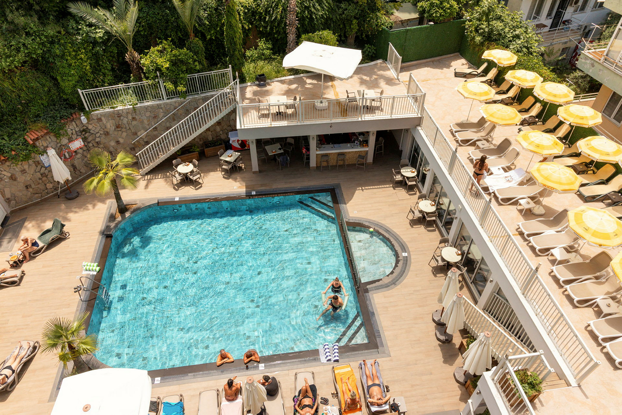 Udendørs pool med solterrasse, liggestole og parasoller omgivet af vegetation og bygninger. (AI-genereret tekst)
