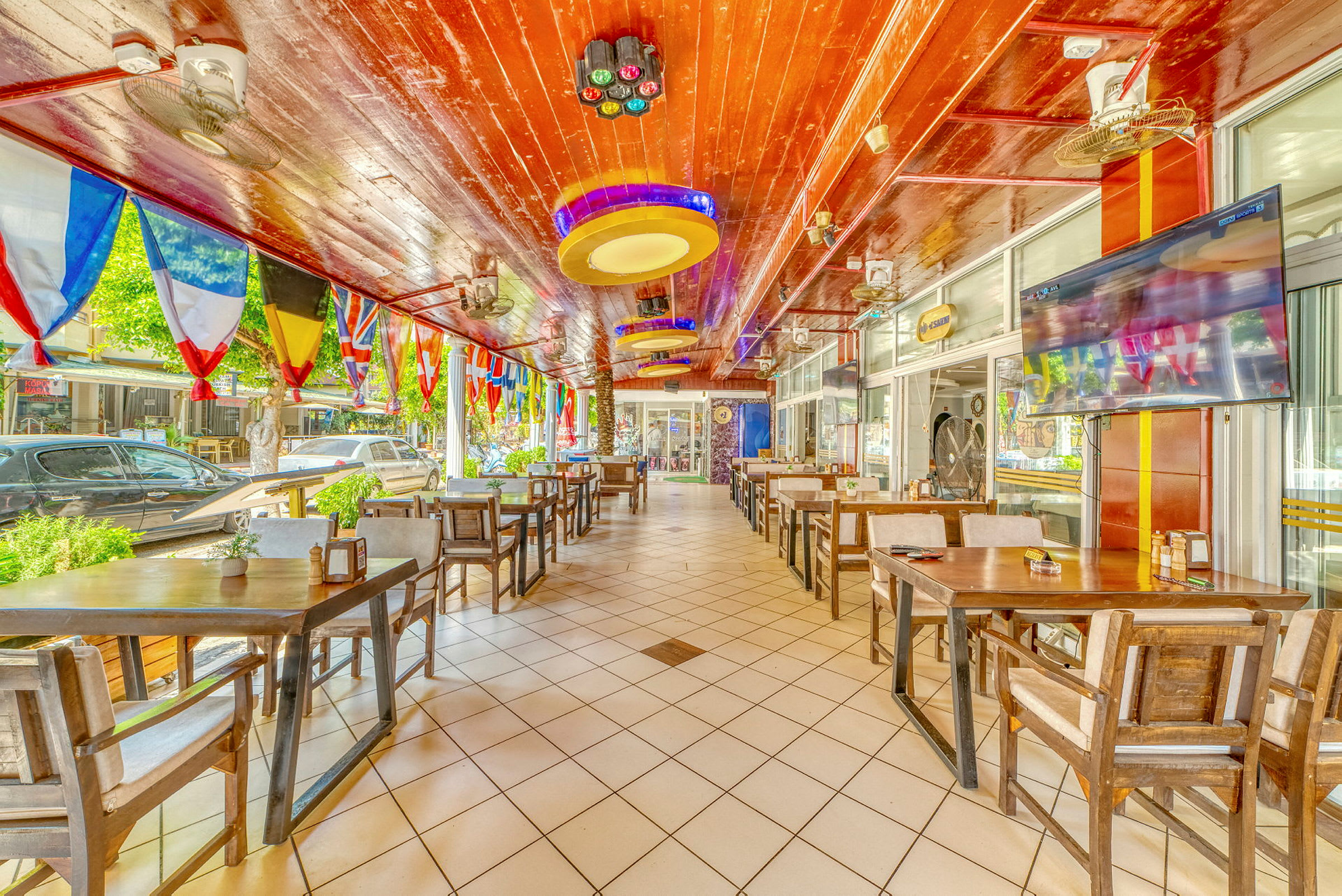 Restaurant med træborde og stole samt farverige flag hængende fra loftet. (AI-genereret tekst)