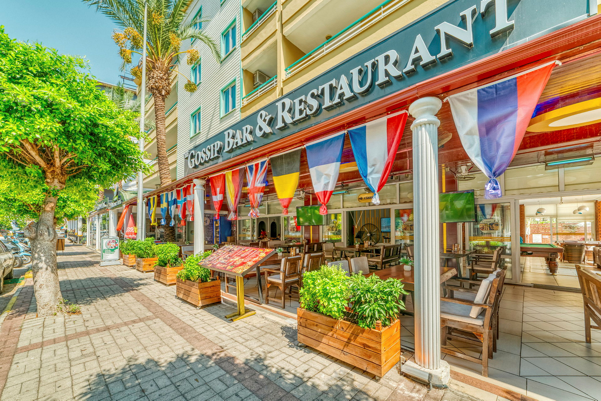 En café eller restaurant med udendørs servering, dekoreret med flag og omgivet af træer og grønt. (AI-genereret tekst)