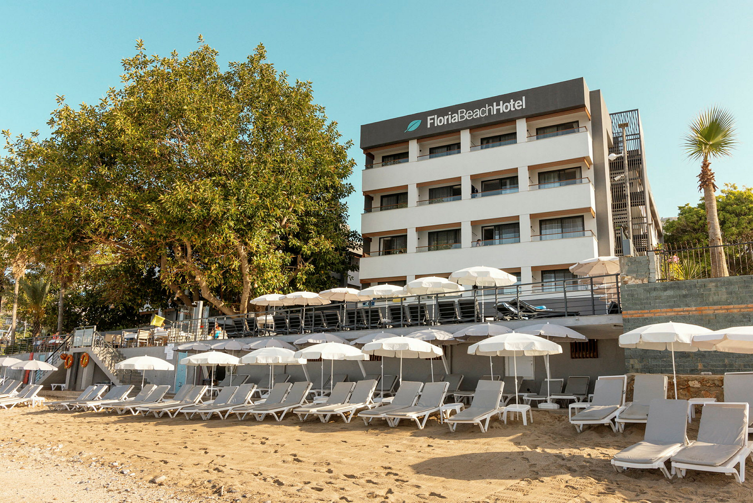 Hvid bygning med skiltet 'Floria Beach Hotel' og en sandstrand med liggestole og parasoller. (AI-genereret tekst)