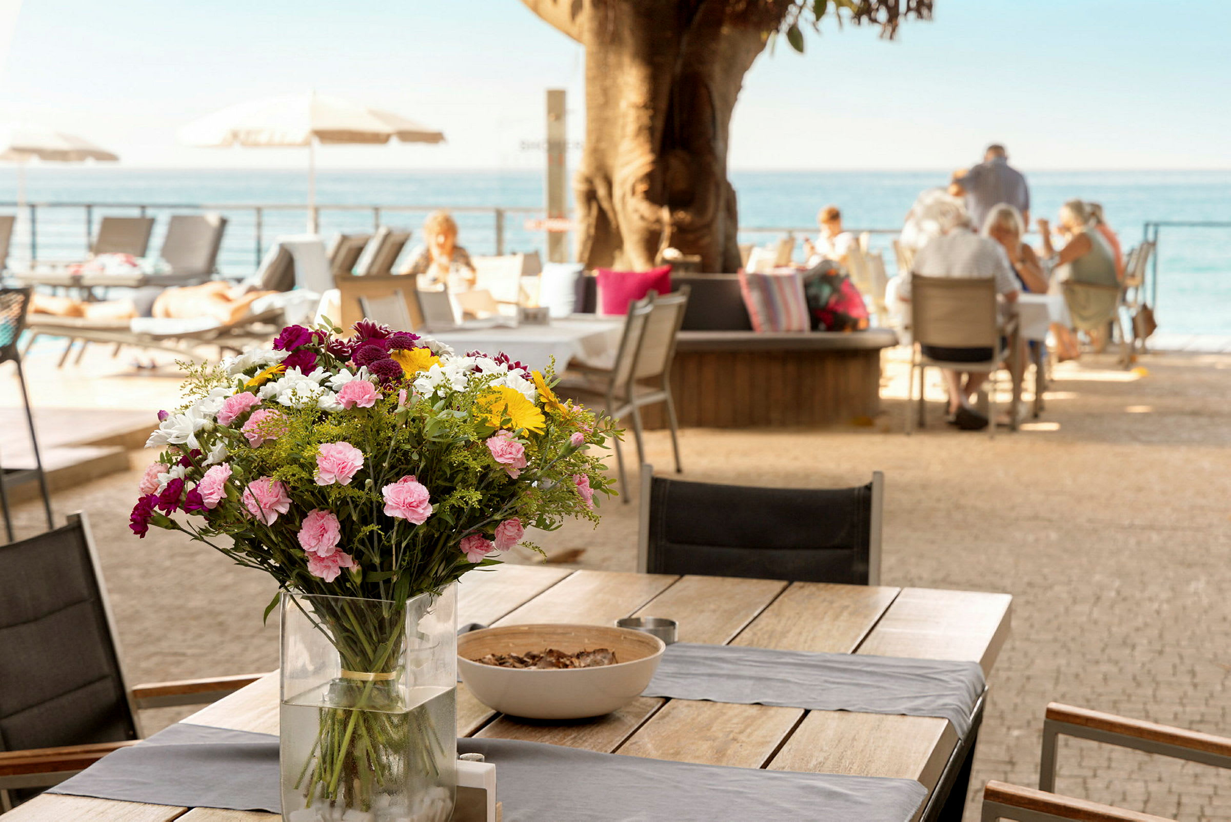 Et bord med en vase fyldt med farverige blomster, med mennesker der sidder ved andre borde i baggrunden tæt på havet. (AI-genereret tekst)