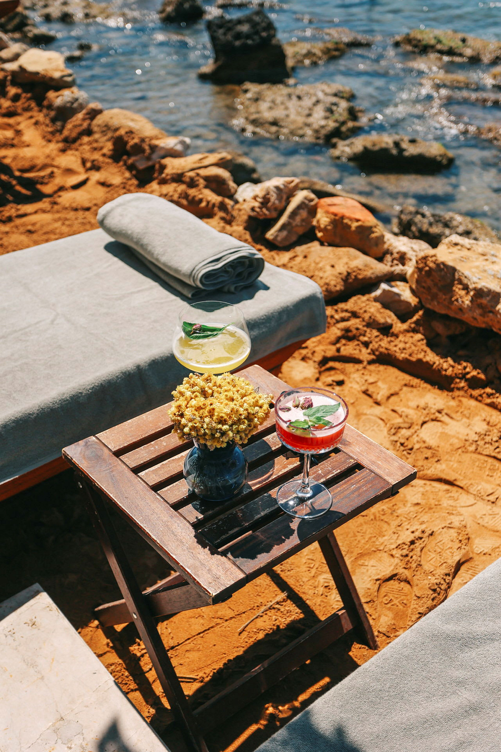 En strandscene med et foldbart træbord med to dekorative drinks og en blomsterkrukke. Baggrunden viser havvand og stenede kyst. (AI-genereret tekst)