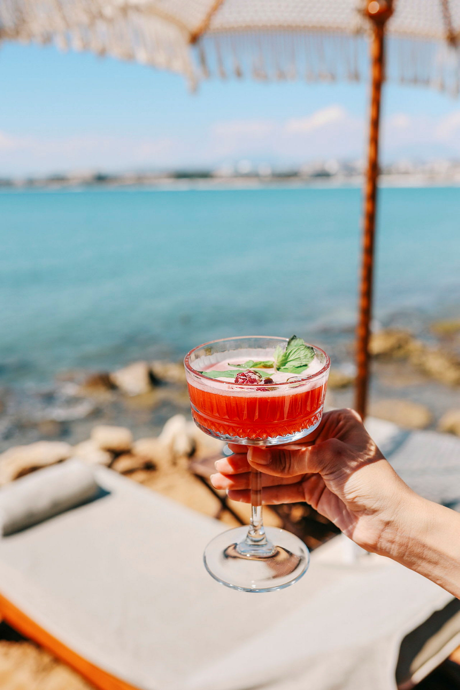 Hånd holder et glas drink med rødfarvet drik og pynt ved strandmiljø. (AI-genereret tekst)