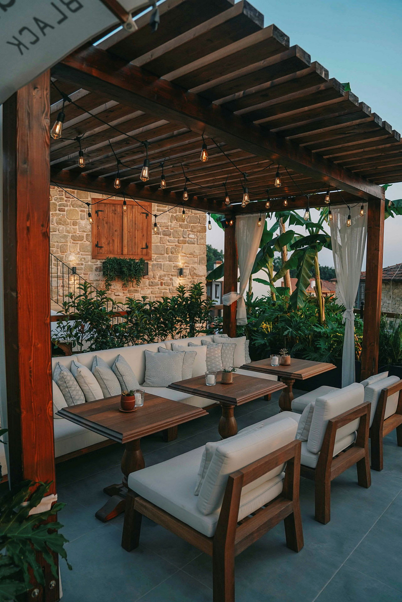Udendørs lounge med træborde og hvide puder under pergola. (AI-genereret tekst)