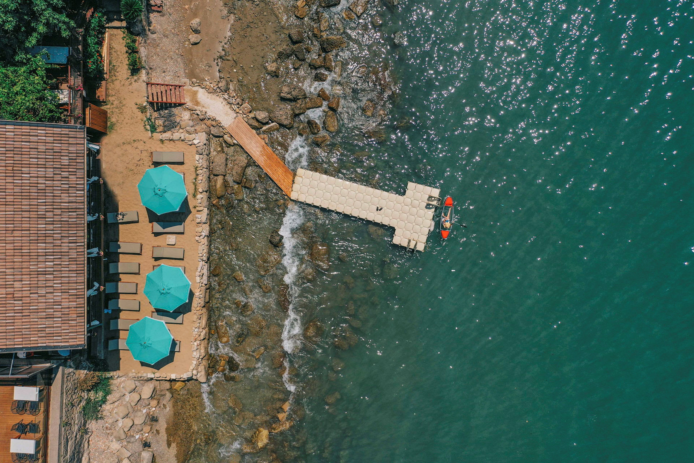 Luftfoto af en badebro med en enkelt kajak i en klarblå sø, omgivet af en strand med liggestole og parasoller. (AI-genereret tekst)