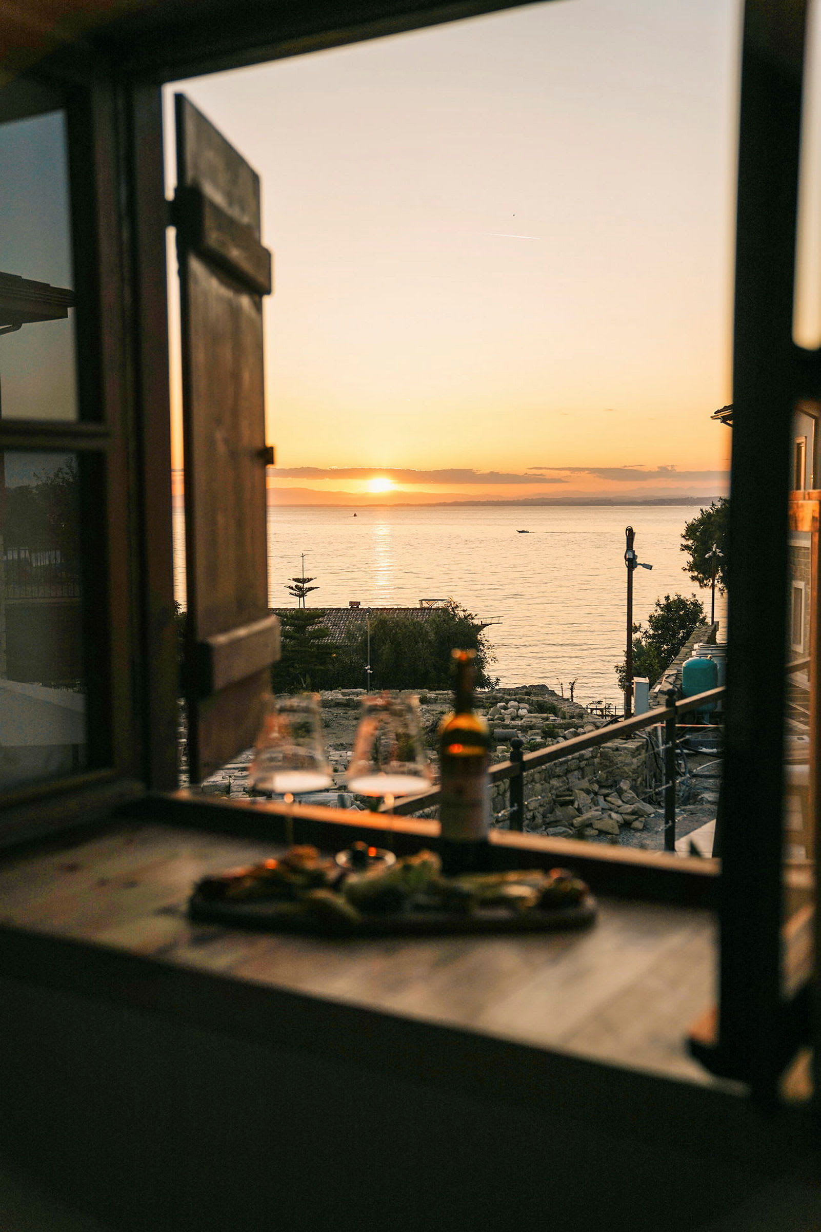 Udsigt fra et vindue mod en solnedgang over havet, med glas og en flaske på en bænk. (AI-genereret tekst)
