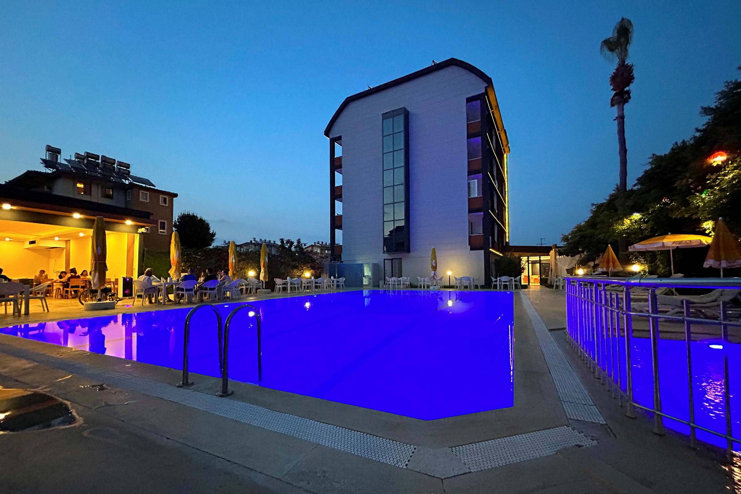 En hotelbygning med en oplyst pool foran, omgivet af stole og parasoller ved skumring. (AI-genereret tekst)