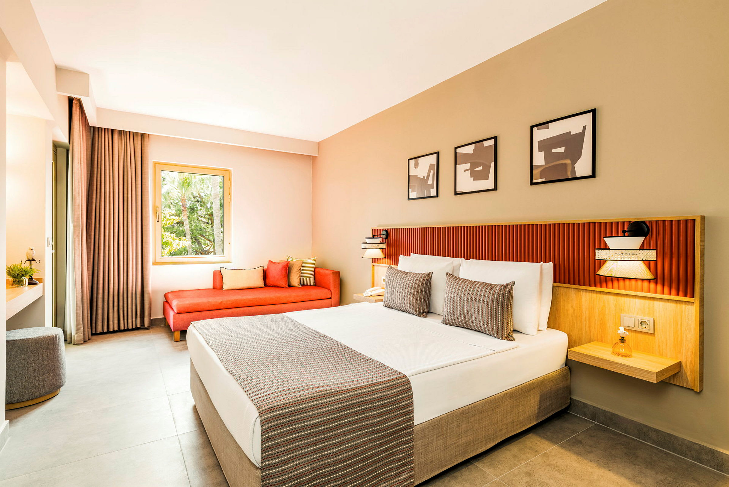 Et moderne hotelværelse med en bred seng med hvide lagner og dekorative puder. Der er en rød sofa ved vinduet og tre kunstværker på væggen over sengen. (AI-genereret tekst)