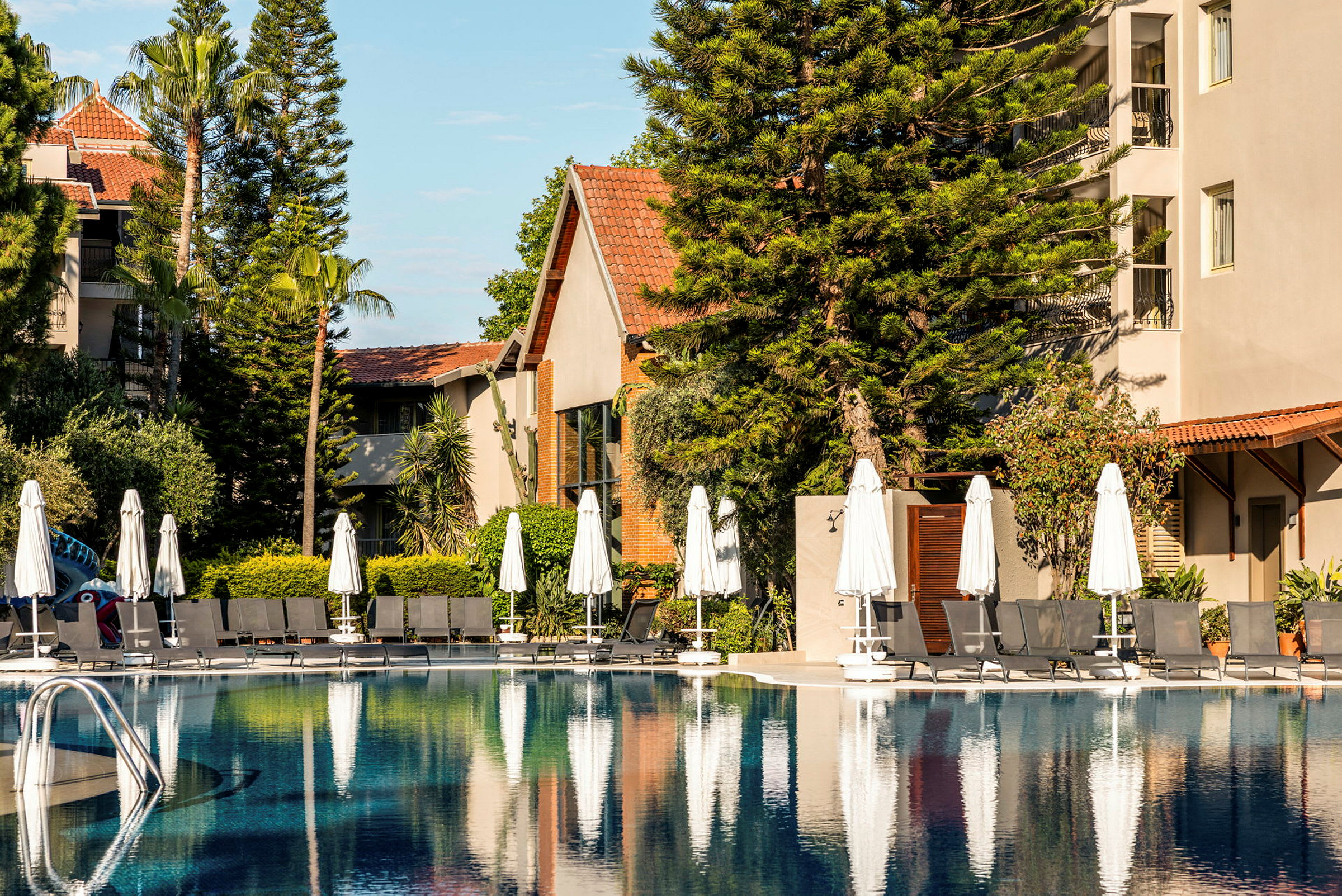 Billede af en pool ved et hotel, omgivet af liggestole, parasoller og frodig beplantning. (AI-genereret tekst)