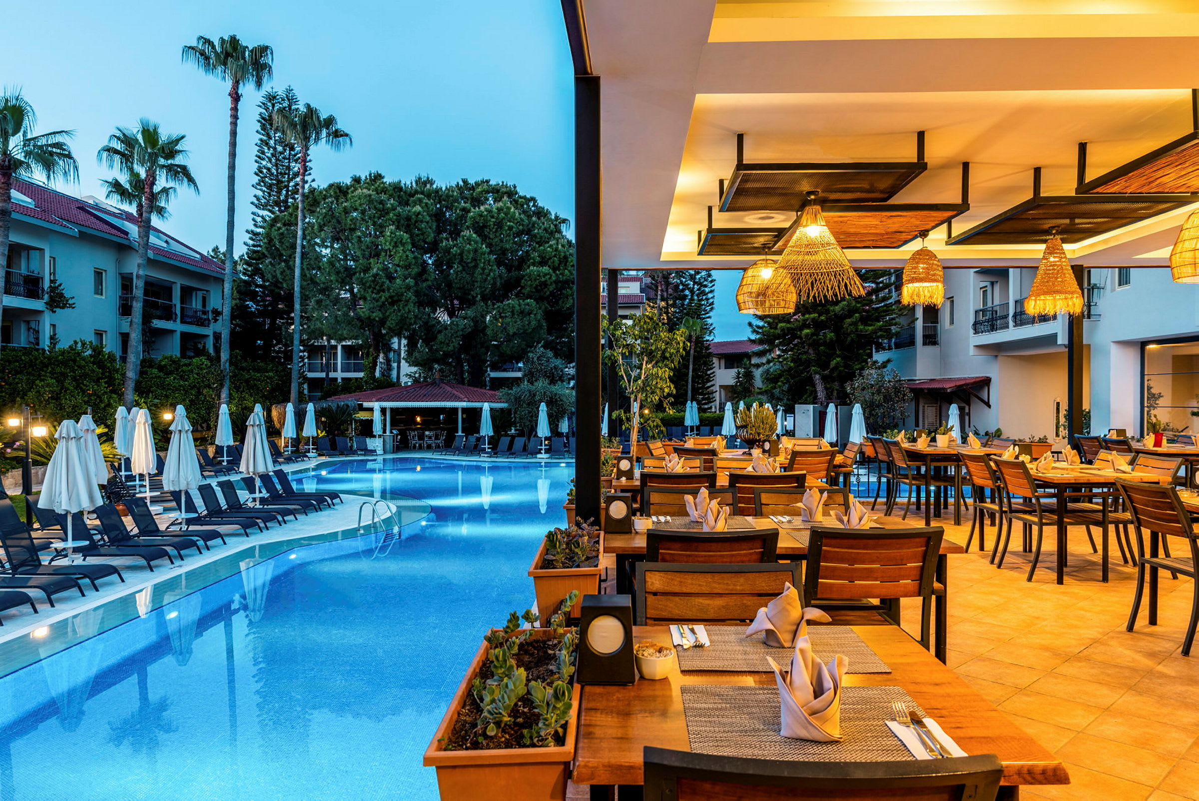 En udendørsrestaurant med borde dækket til middag ved siden af en pool omgivet af palmer. (AI-genereret tekst)